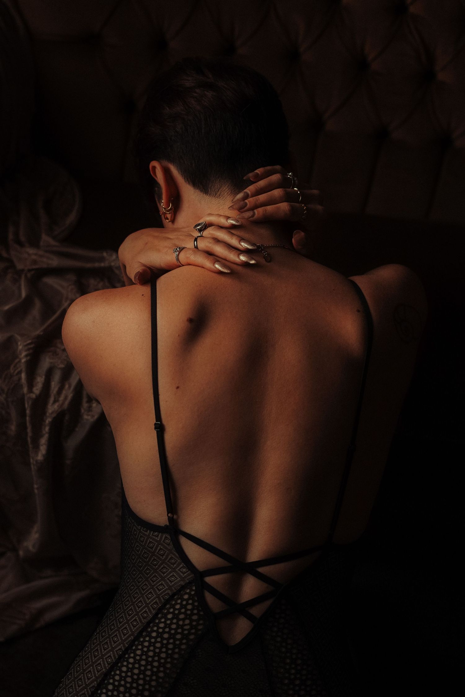 Back view of a woman in a strappy low-back bodysuit with hands on neck against tufted headboard.