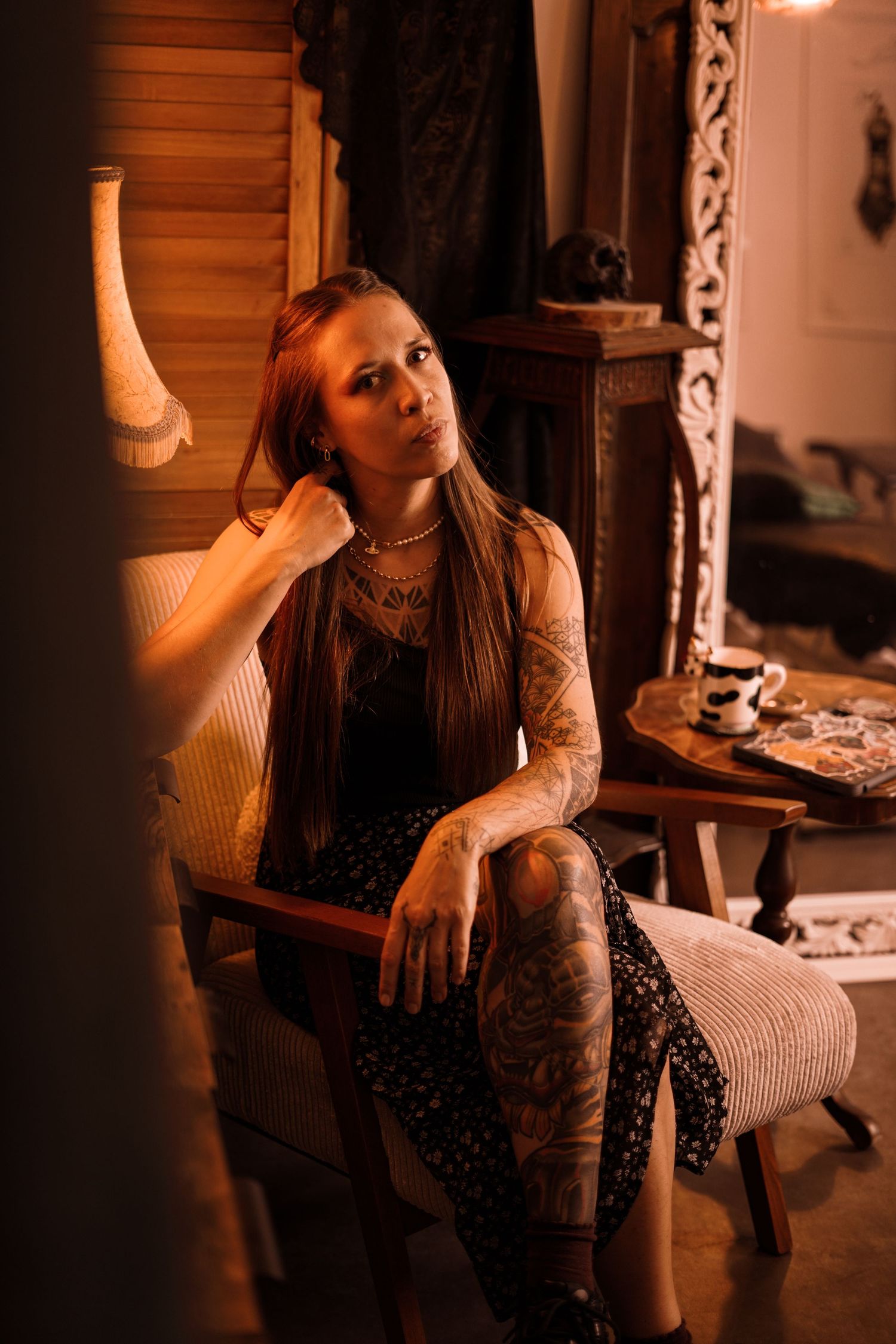 Tattooed woman with long hair sitting in a chair in a warmly lit vintage-style room.