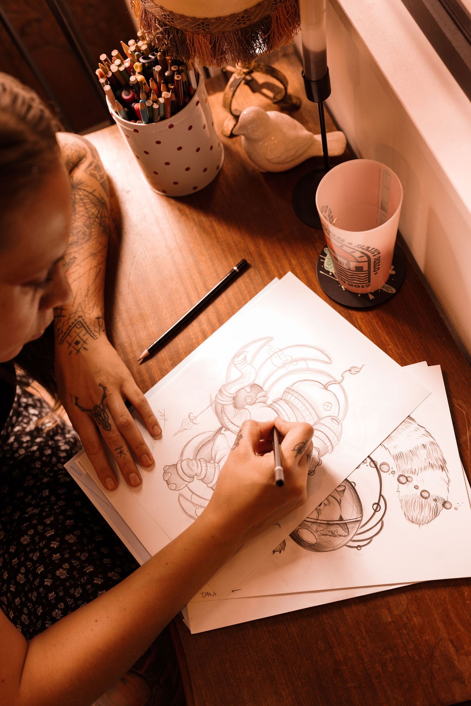 Tattooed artist sketching detailed illustrations at a wooden desk with pencils and warm lighting.
