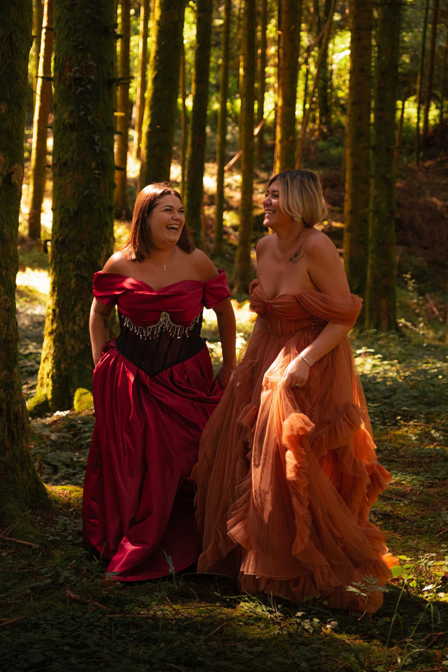 Two women in elegant red and orange gowns laughing in a sunlit forest setting.