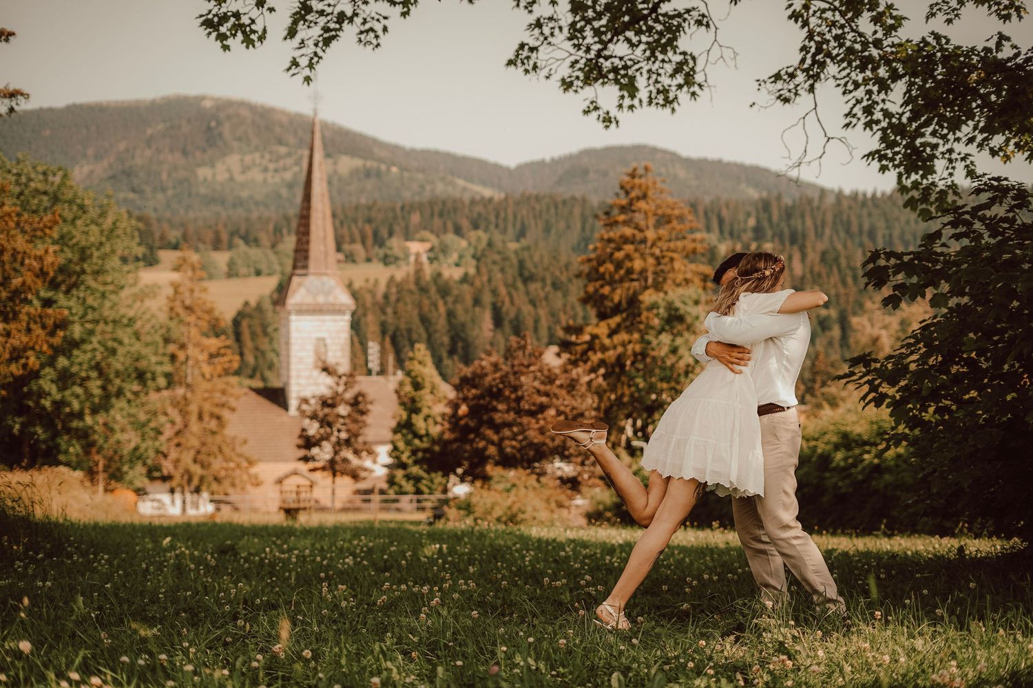 Bride and groom embrace and spin in a meadow with a picturesque church and mountains behind.