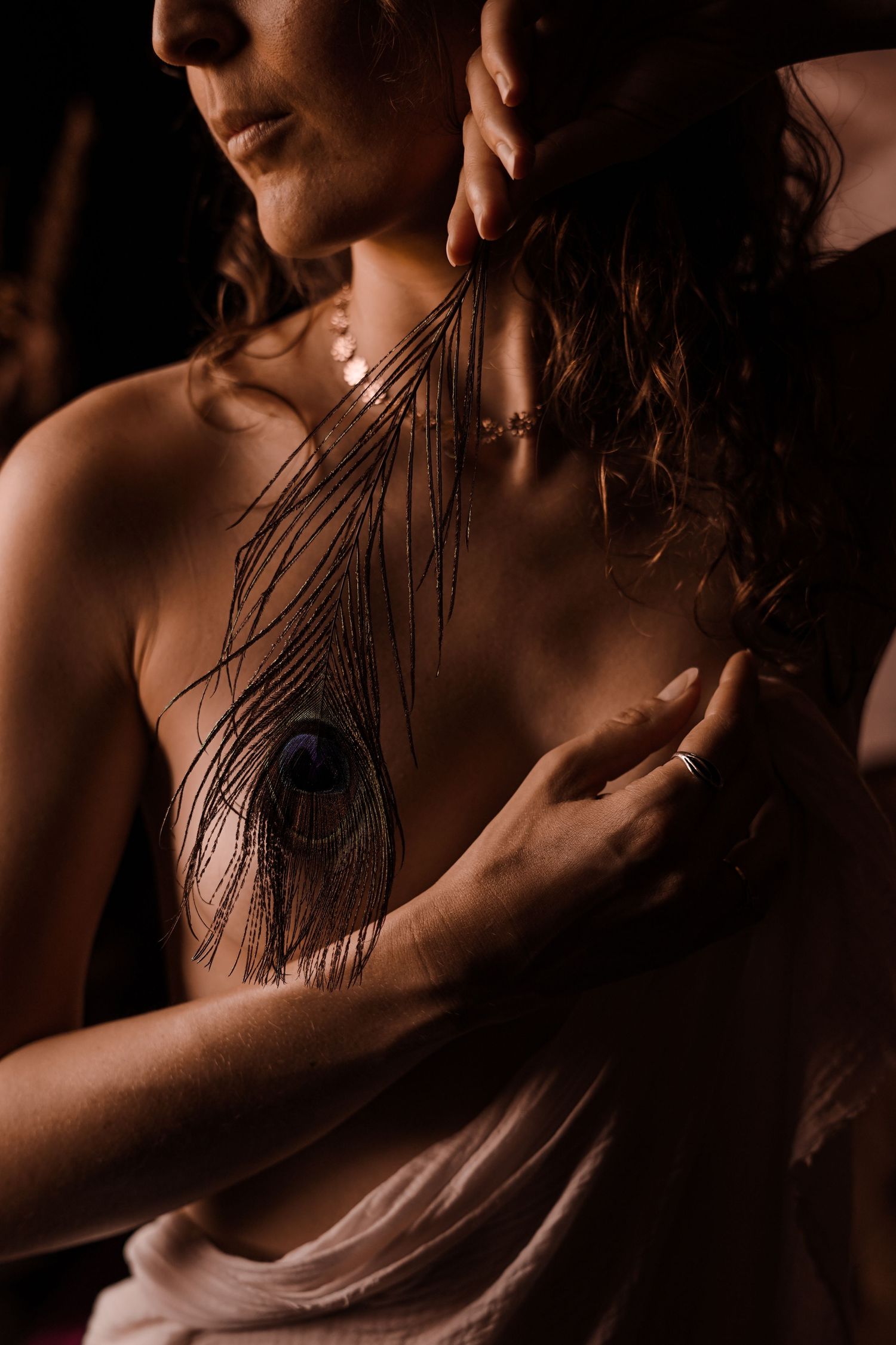 Woman holding peacock feather against chest while being embraced in warm moody dark lighting.