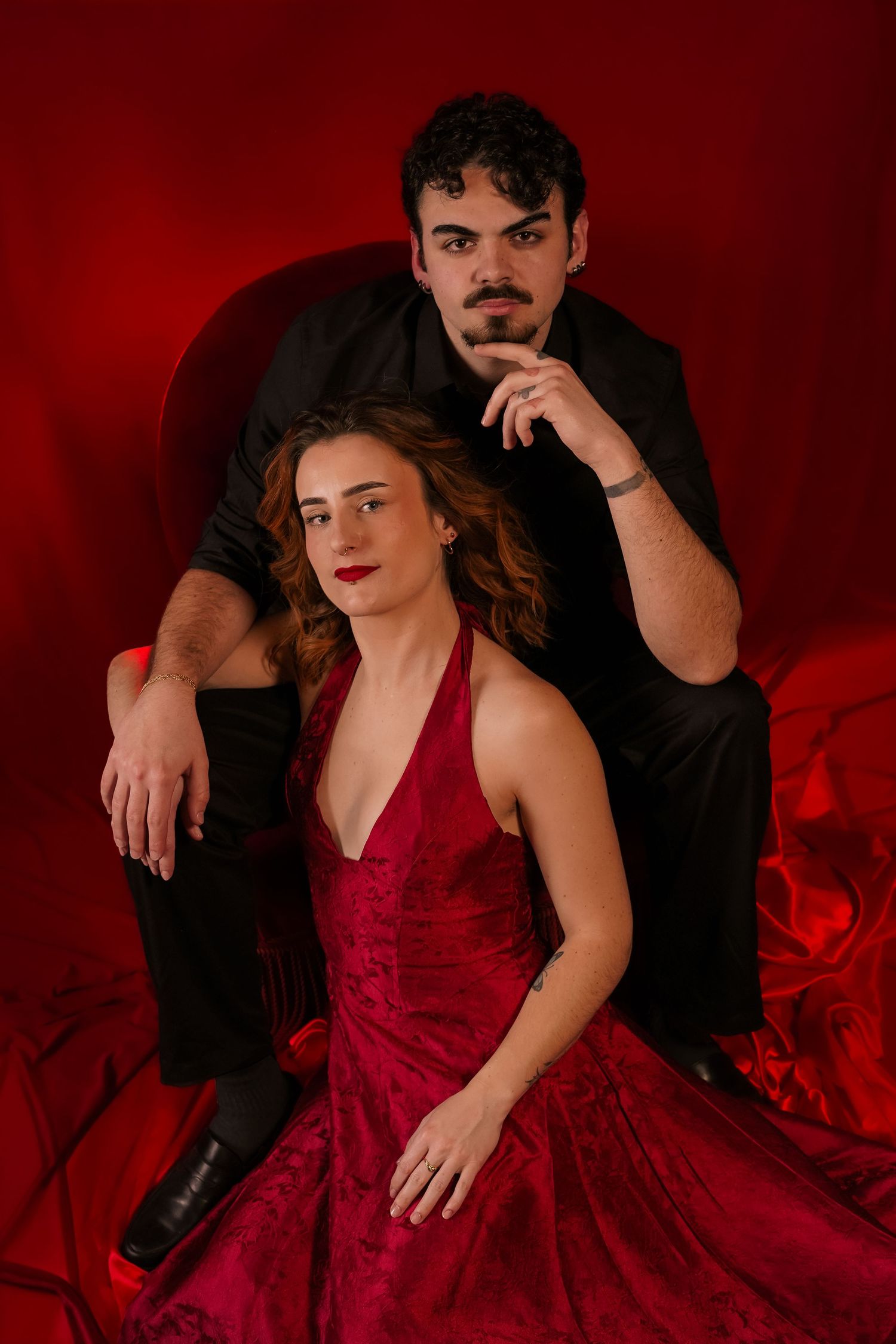 Couple posing dramatically in red studio, woman in red gown seated before man in black suit.