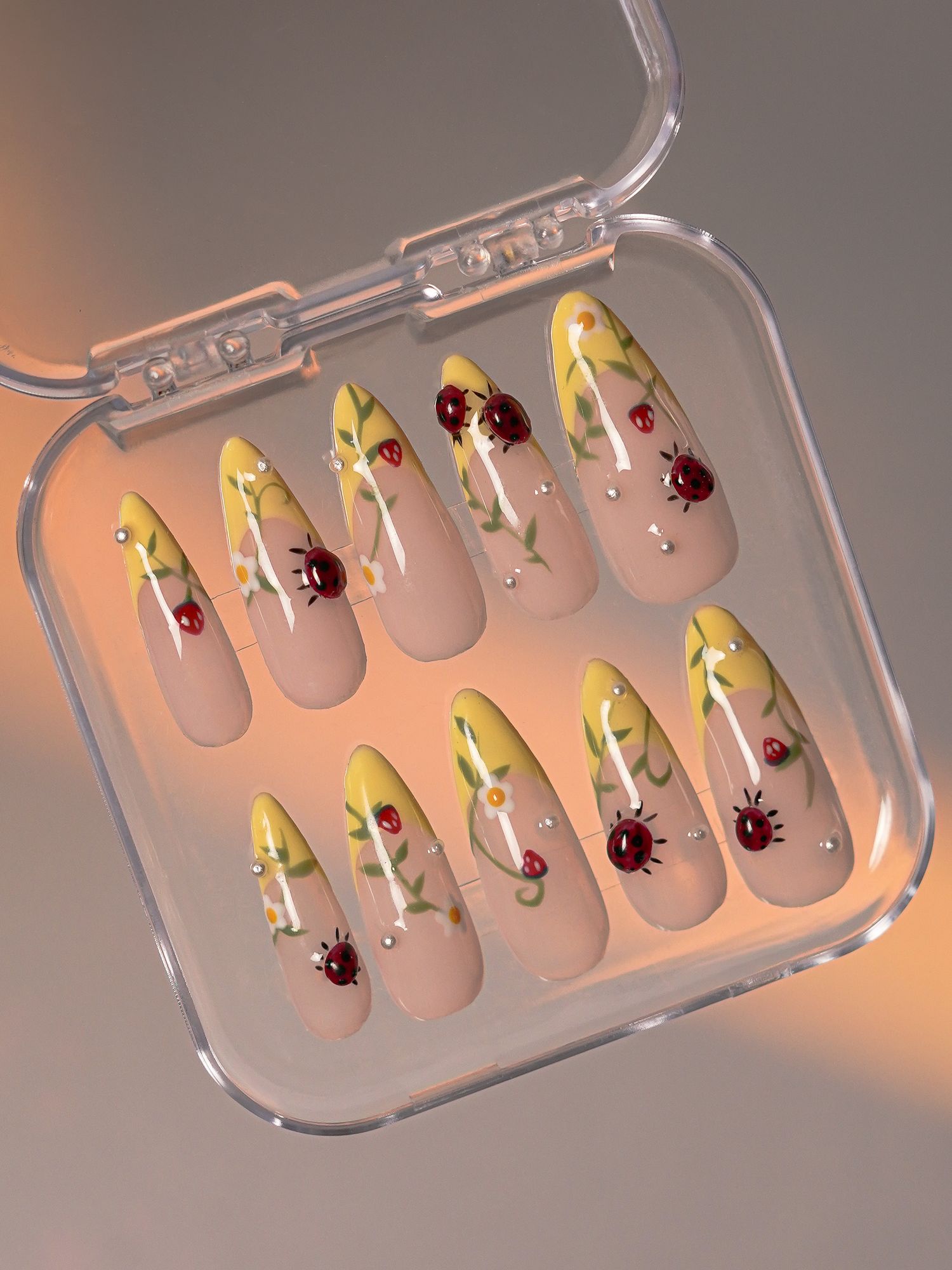Press-on nails in clear case featuring ladybug and floral designs with rhinestones on nude and yellow base.