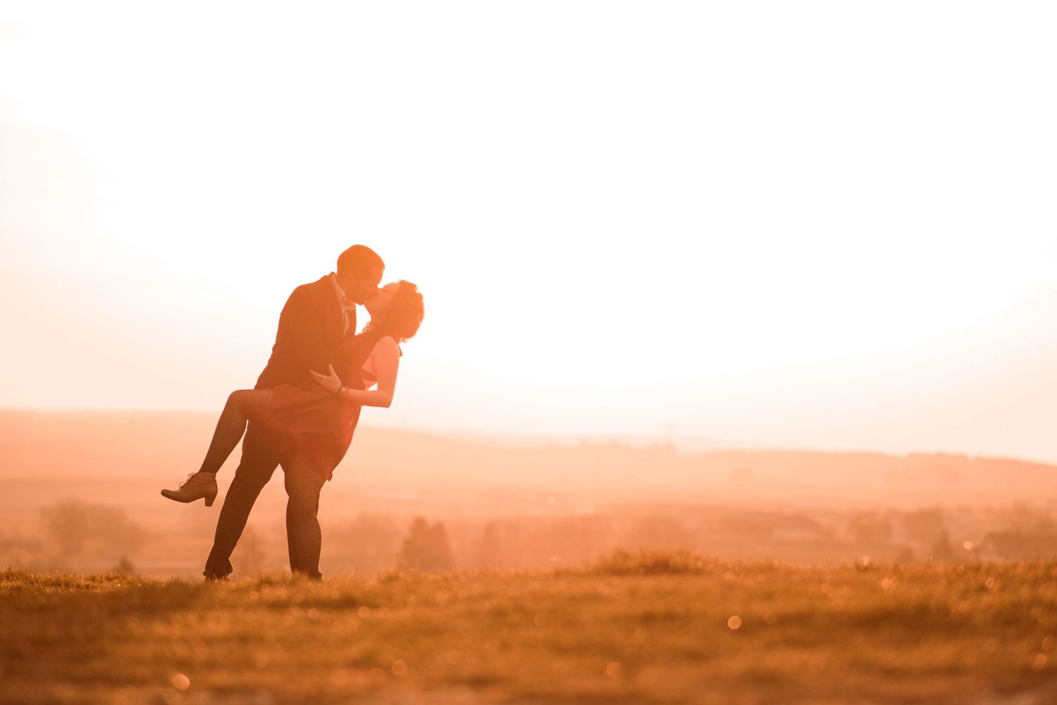 Silhouette of a romantic couple embracing and kissing in a glowing golden sunset field.