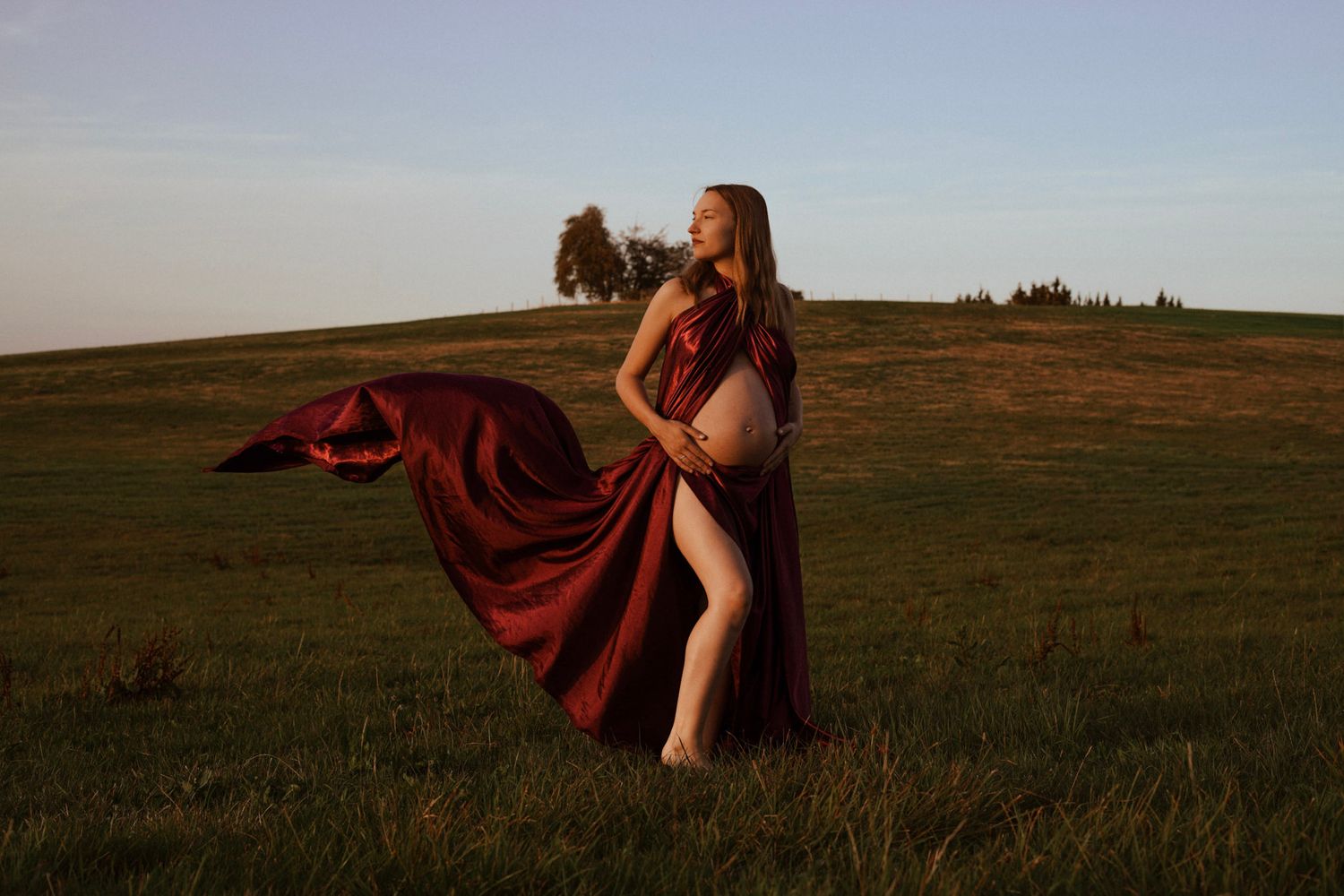 Pregnant woman in flowing red dress standing in green field at golden hour with dramatic fabric movement.