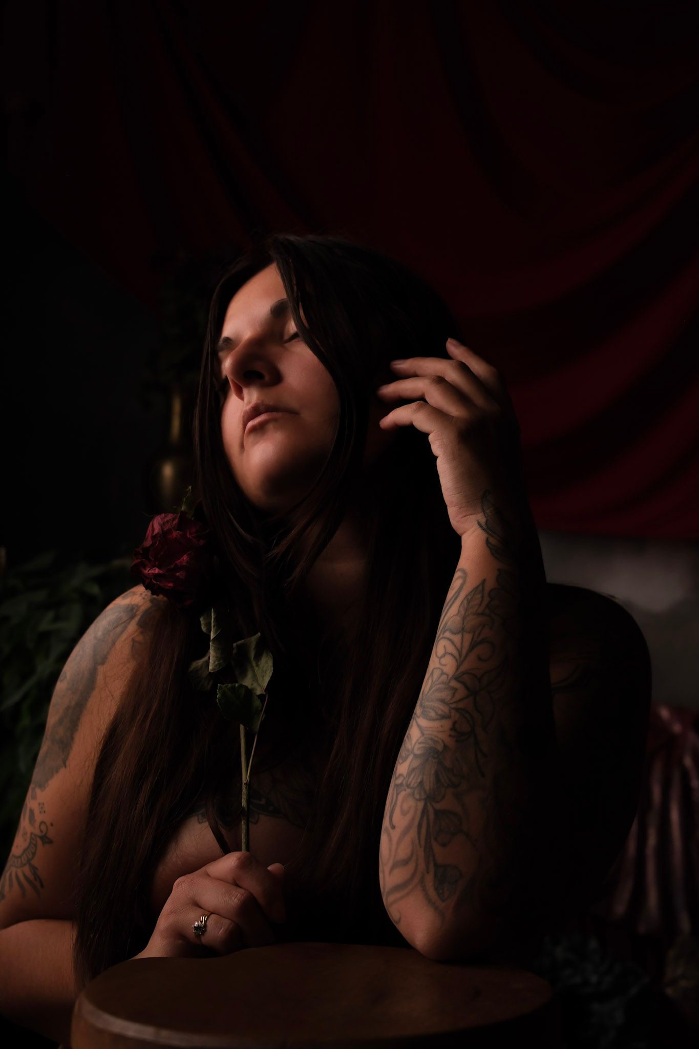 Tattooed woman holding a dark red rose in moody, dramatic lighting with rich red drapery background.