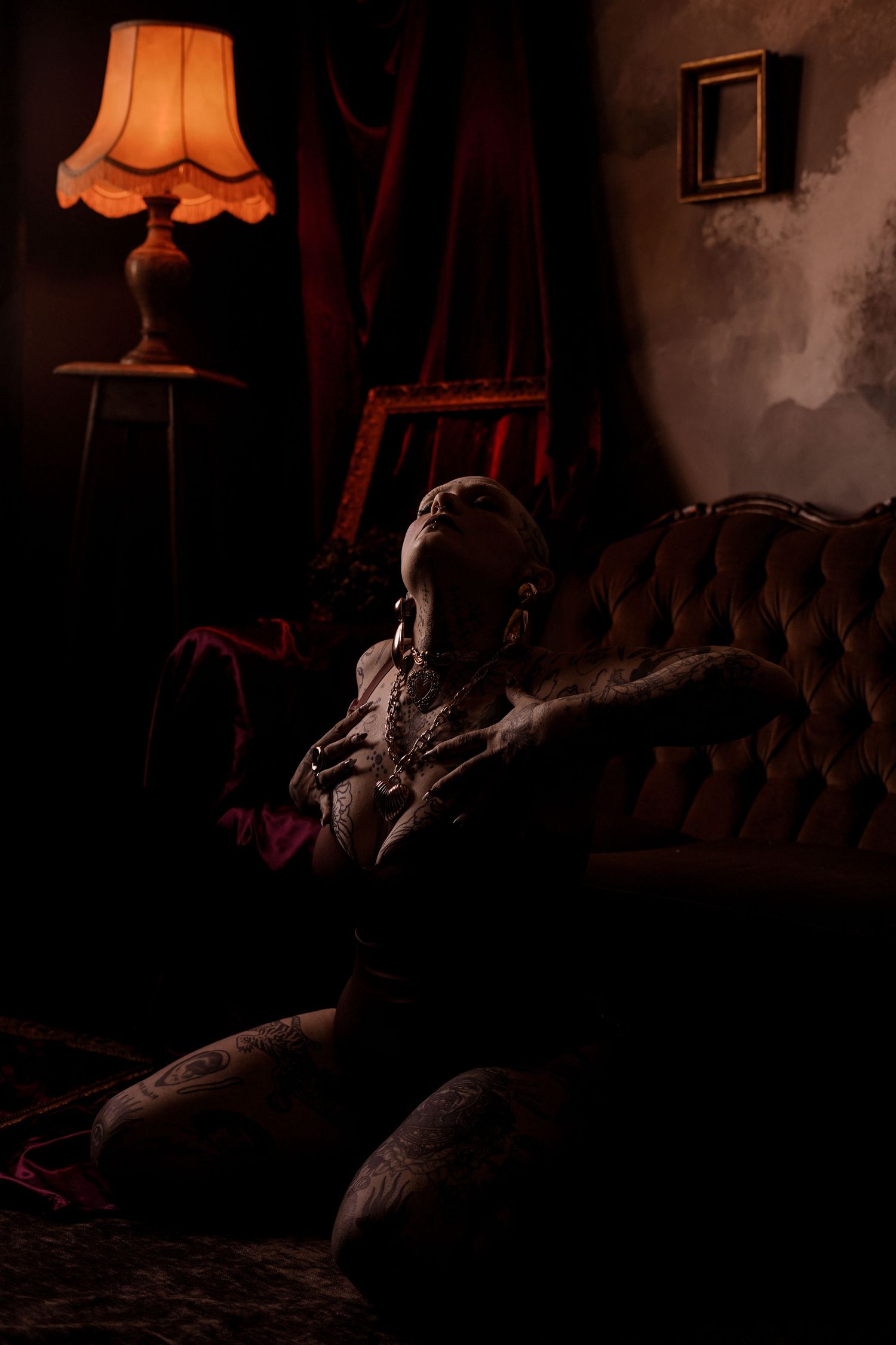 Tattooed person reclining on vintage tufted sofa in dimly lit room with warm amber lamp glow.