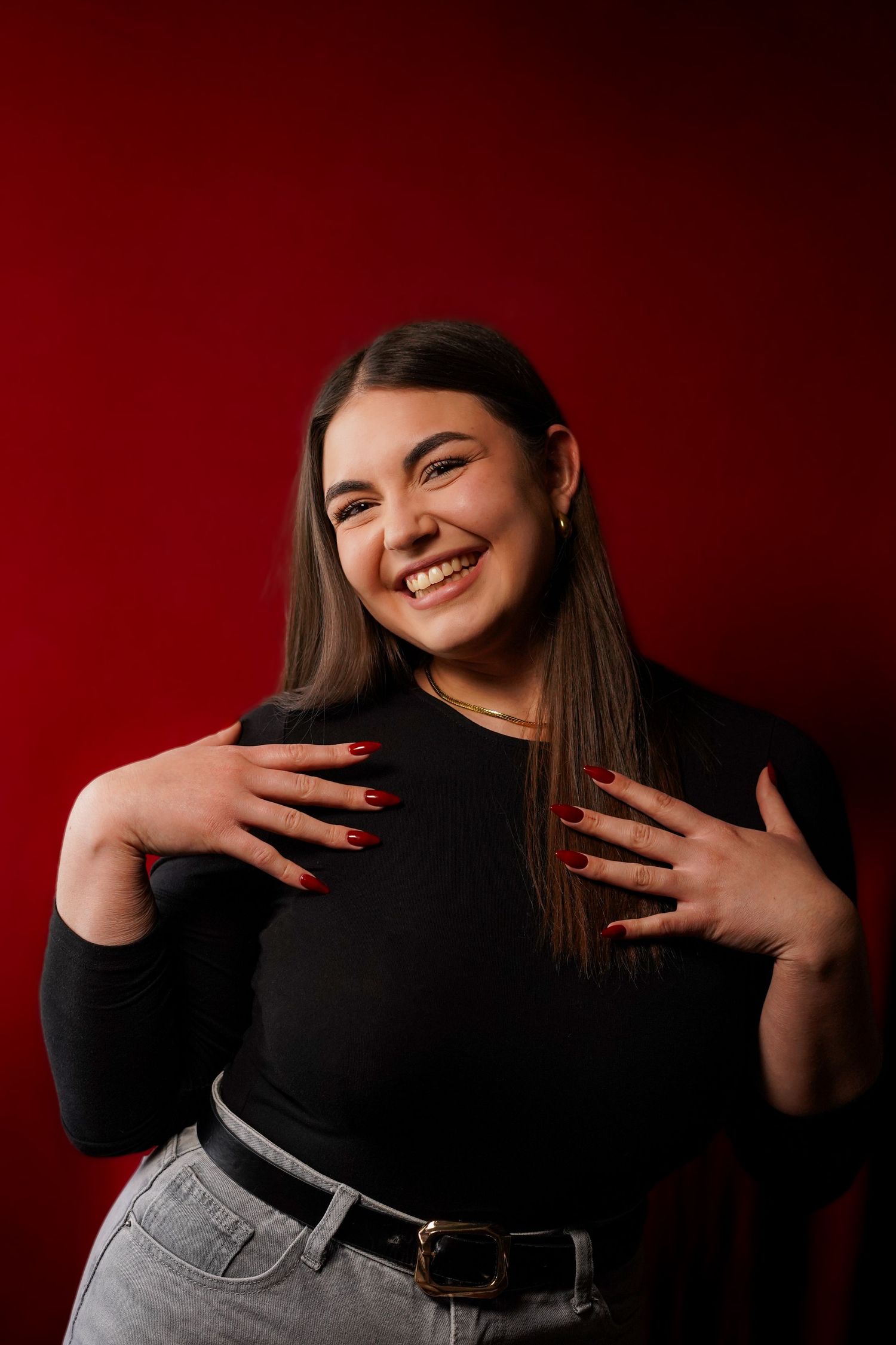Smiling young woman with red nails in black top posing against a red background.