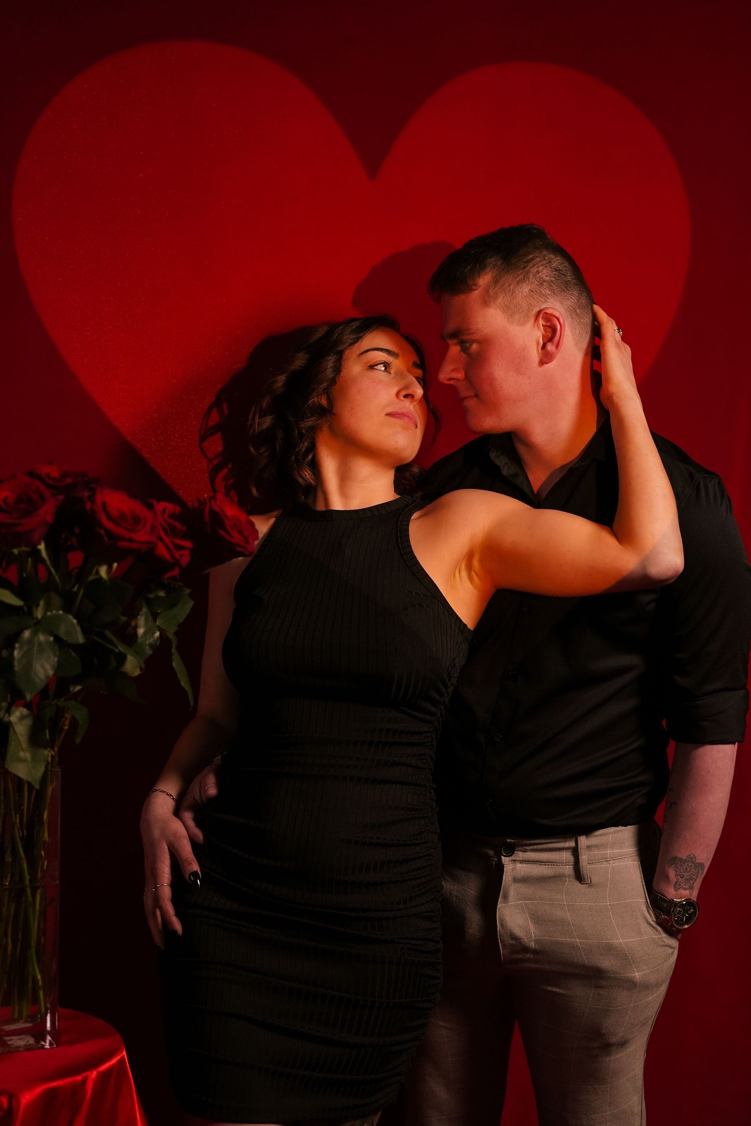 Romantic couple in black outfits embracing before red heart backdrop with roses nearby.
