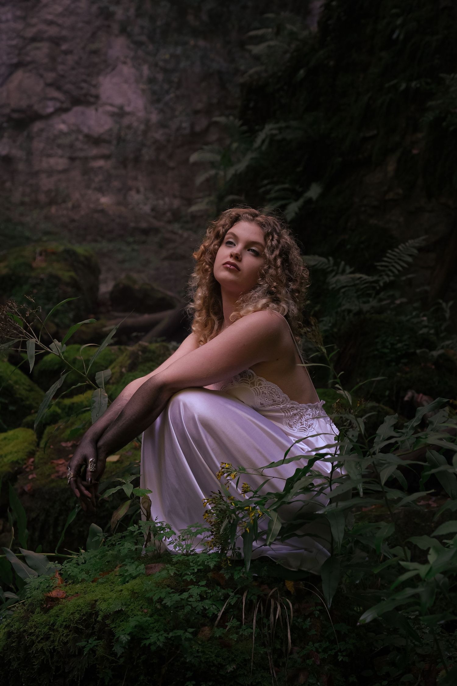 Young woman with curly blonde hair in white lace slip dress sits among ferns in dark rocky forest.