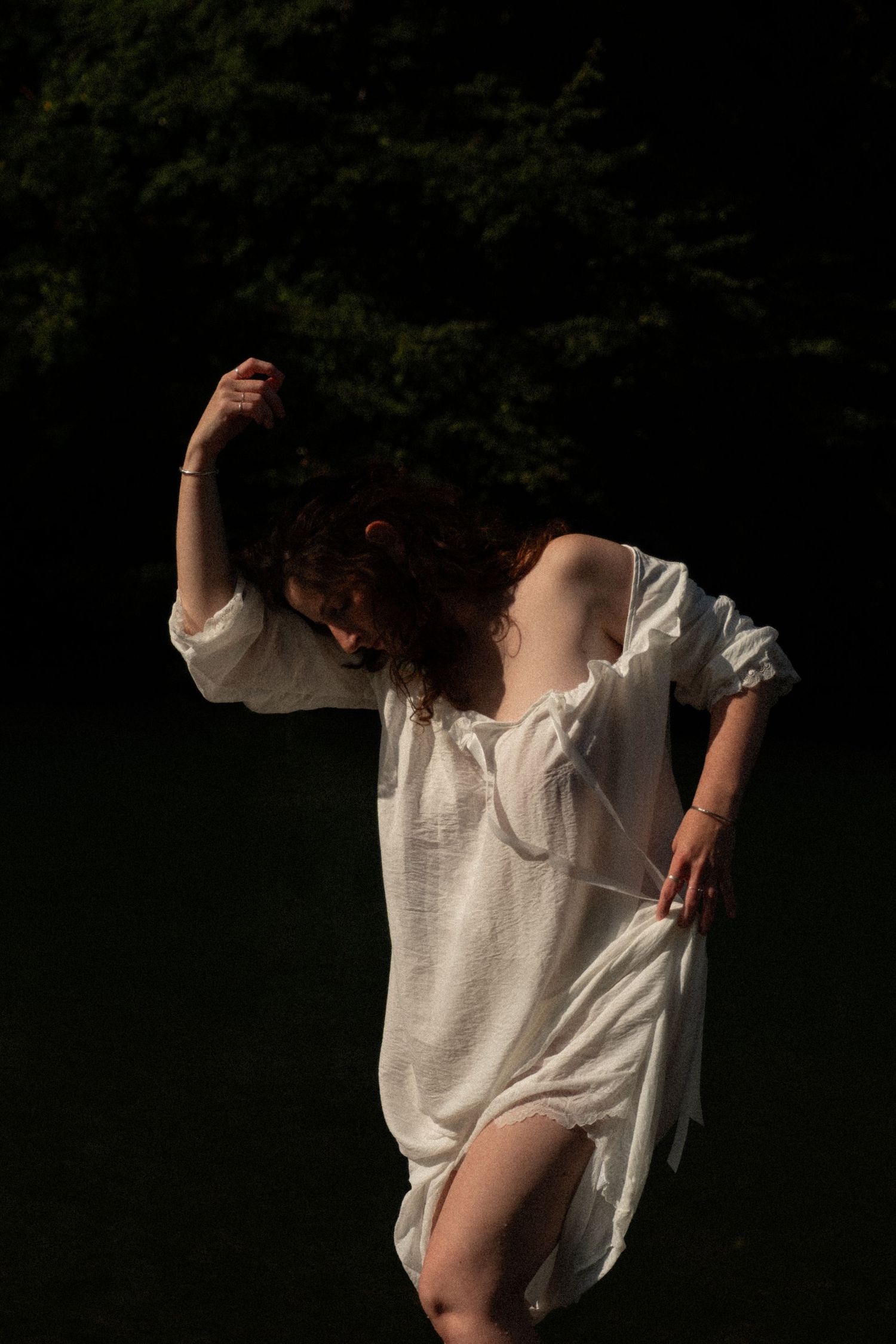 Woman in flowing white linen dress dancing in dramatic light against dark forest backdrop.