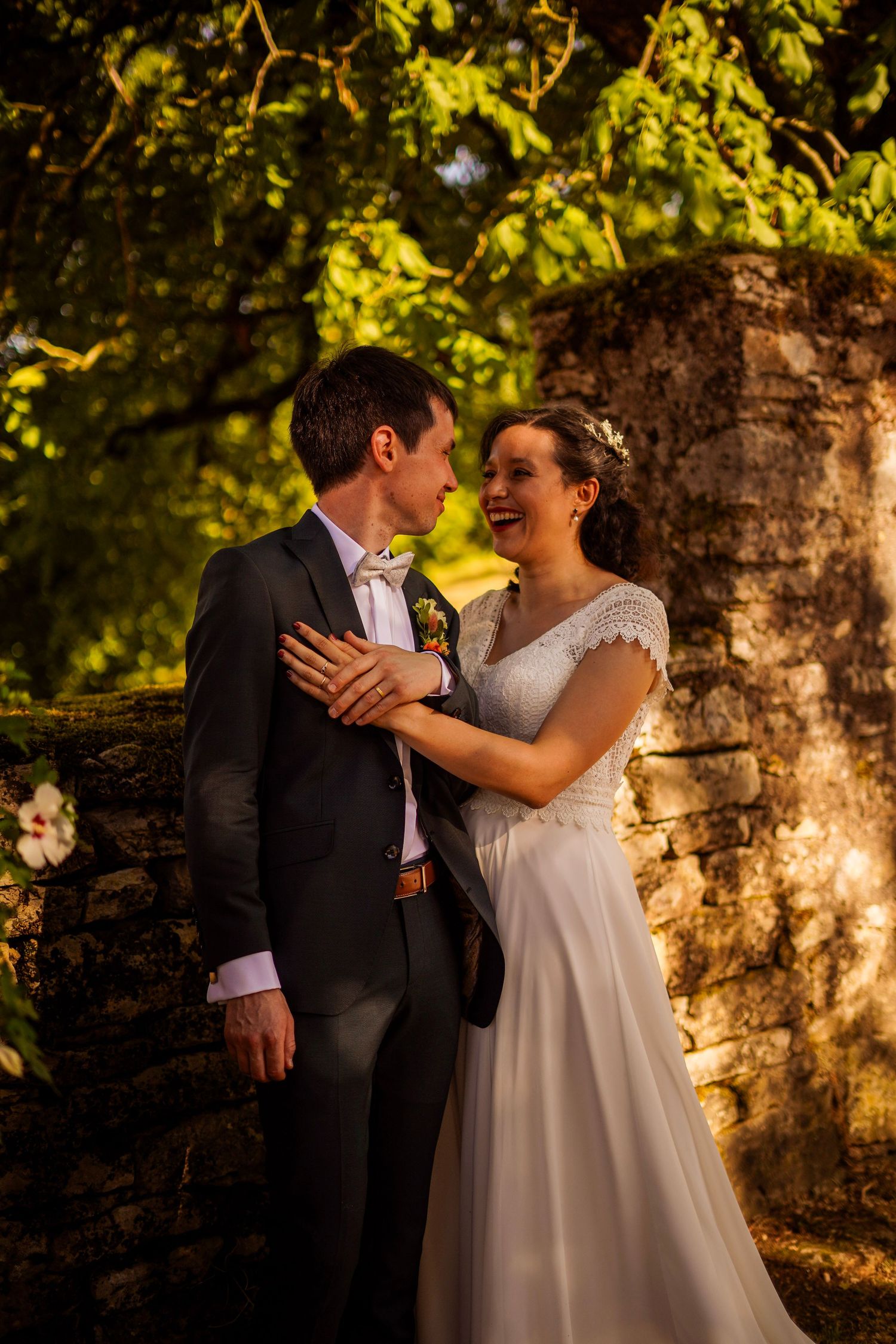 Happy bride and groom embrace near a stone wall surrounded by lush green trees in golden sunlight.