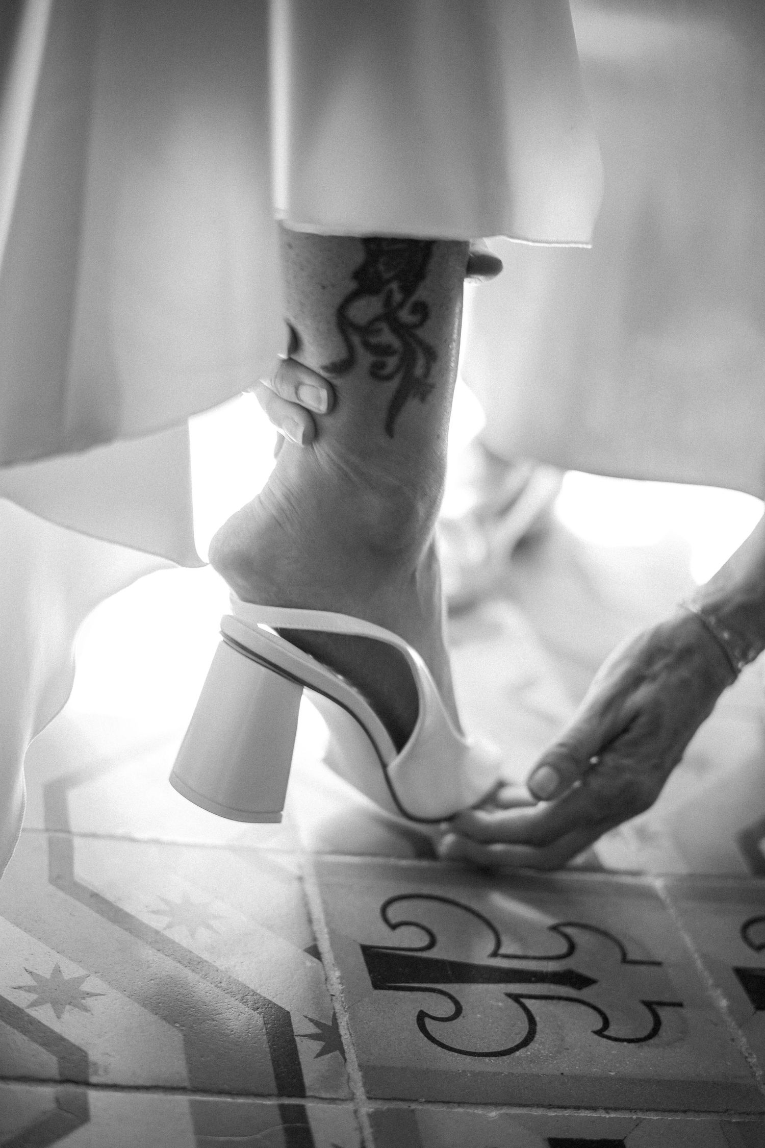 Black and white close-up of bride's tattooed ankle in white block heel wedding shoe.