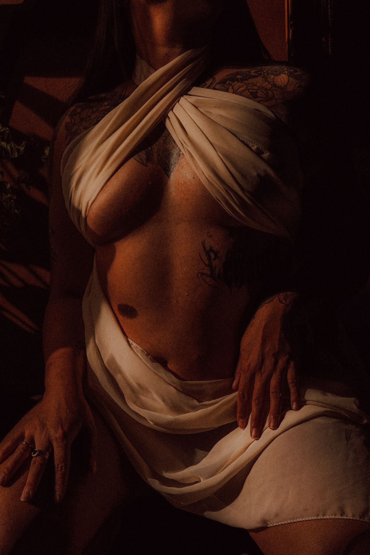 Artistic boudoir image of a tattooed torso wrapped in draped white fabric in warm dark studio lighting.