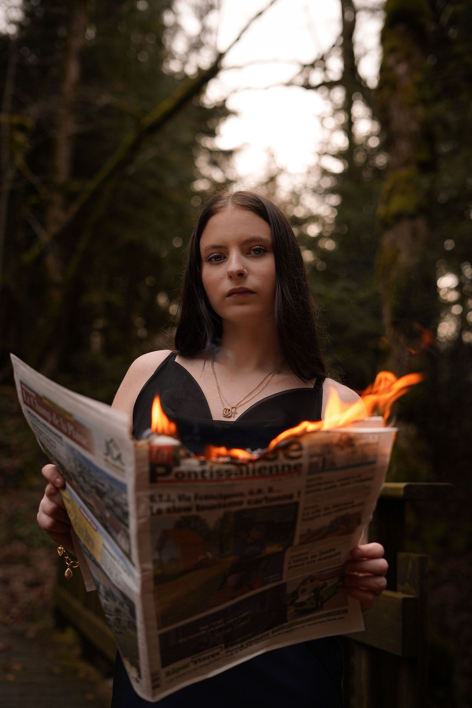 Young woman in black dress reads burning newspaper in forest setting.