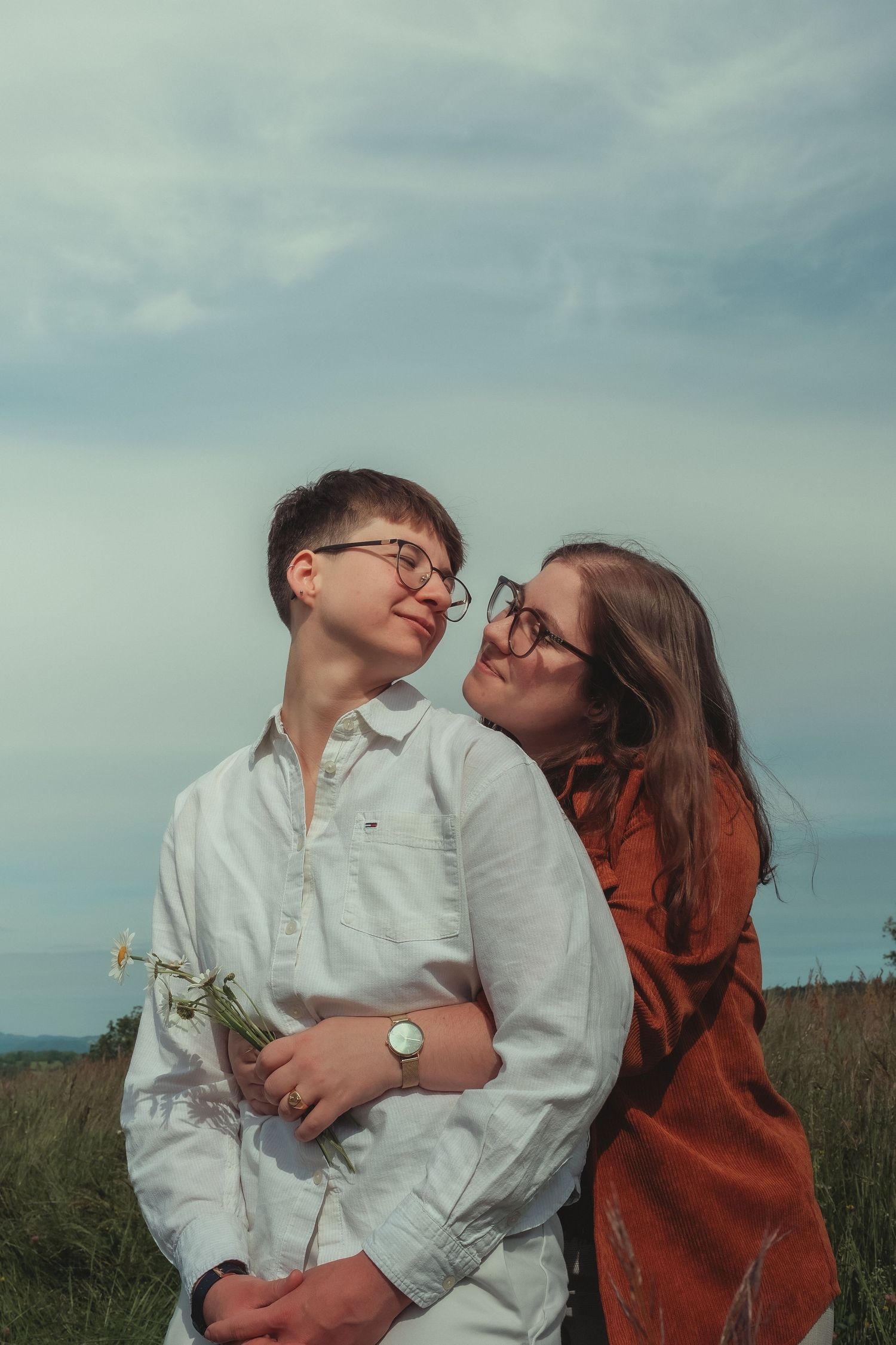 A couple wearing glasses shares a romantic moment in a field, one holding white daisies under cloudy skies.