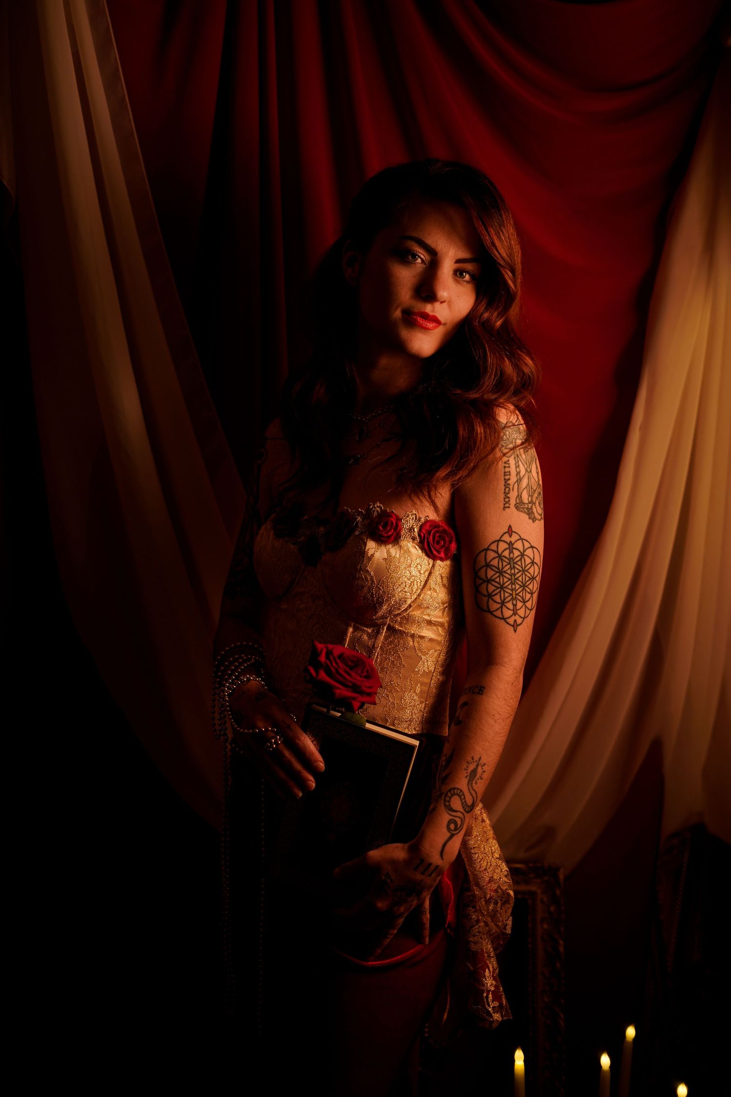 Tattooed woman in corset top poses by candlelight with red drapes.