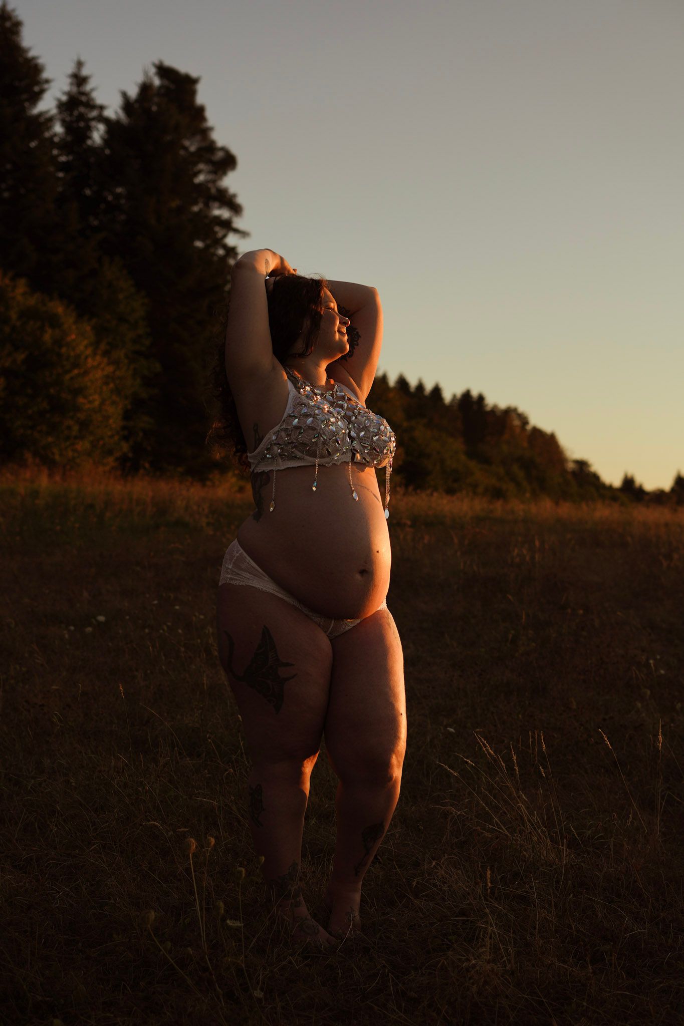 Pregnant woman in bikini posing in golden field at sunset with forest background.