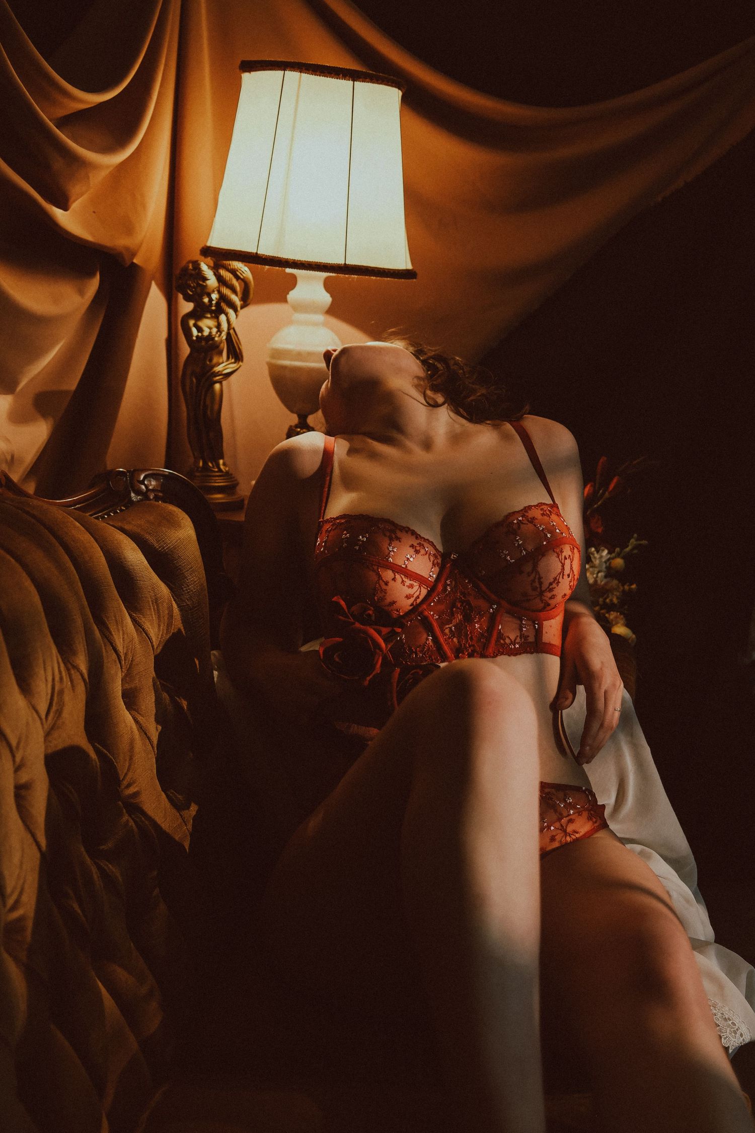 Woman in red lace lingerie reclining on vintage sofa beside antique lamp in warm light.