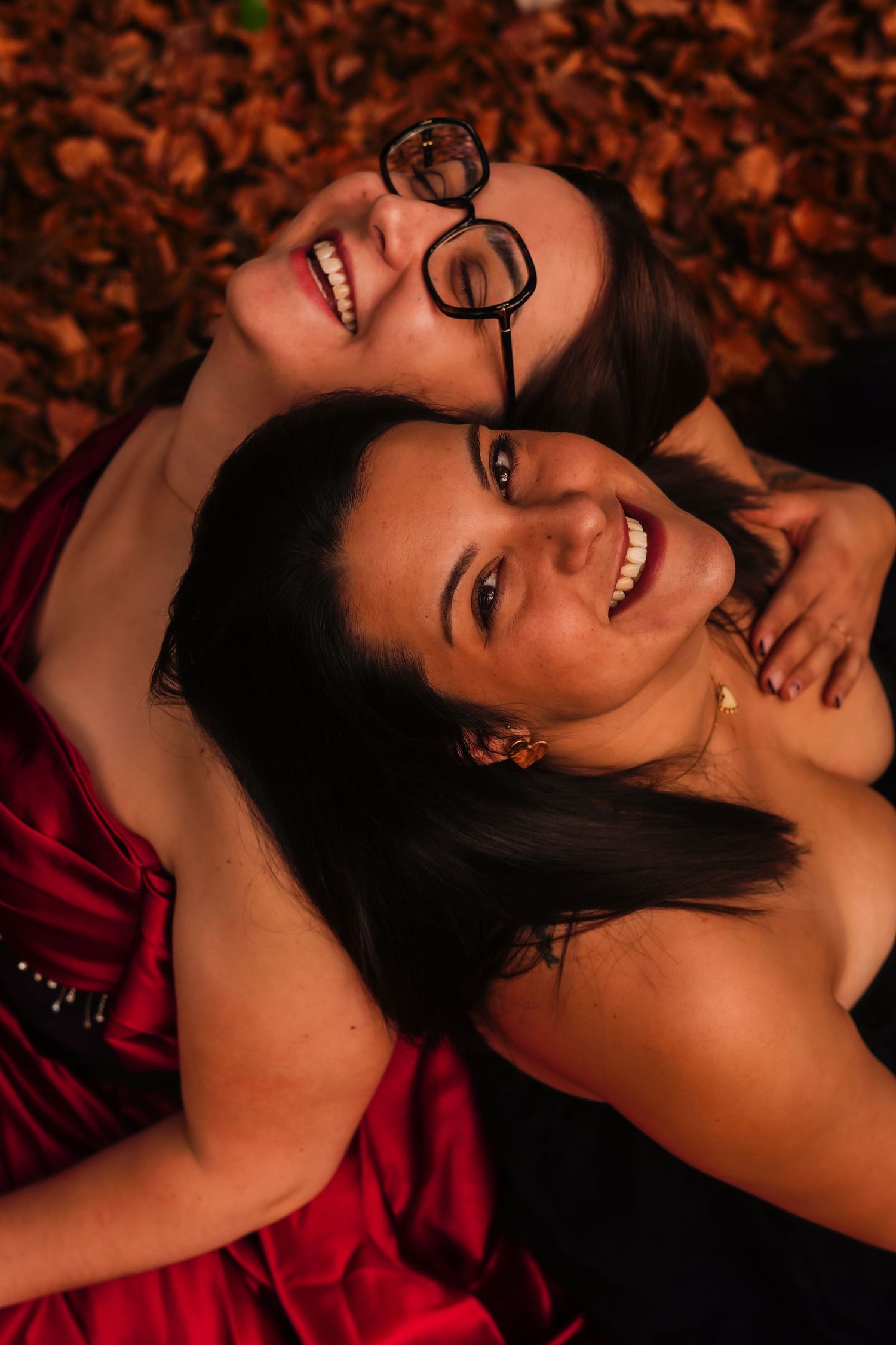 Two smiling women lying on autumn leaves laughing together in joyful outdoor portrait.