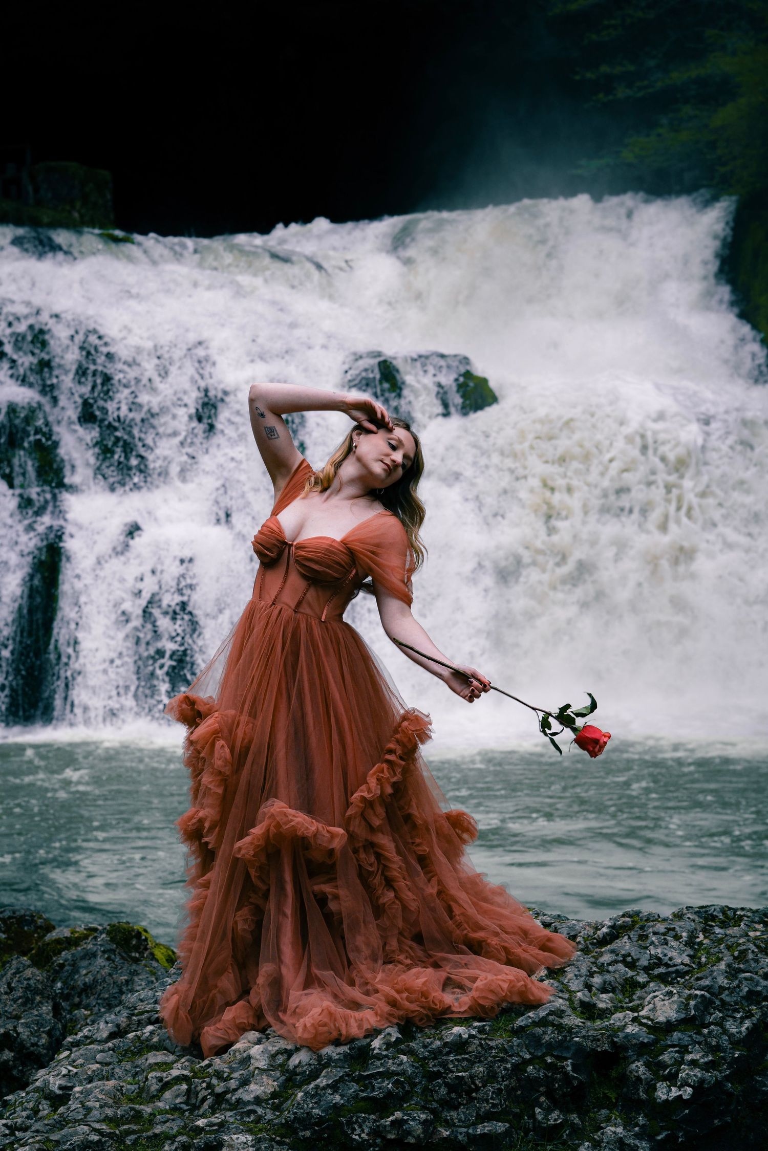 A woman in a rust ruffled tulle gown poses dramatically with a red rose before a cascading waterfall.