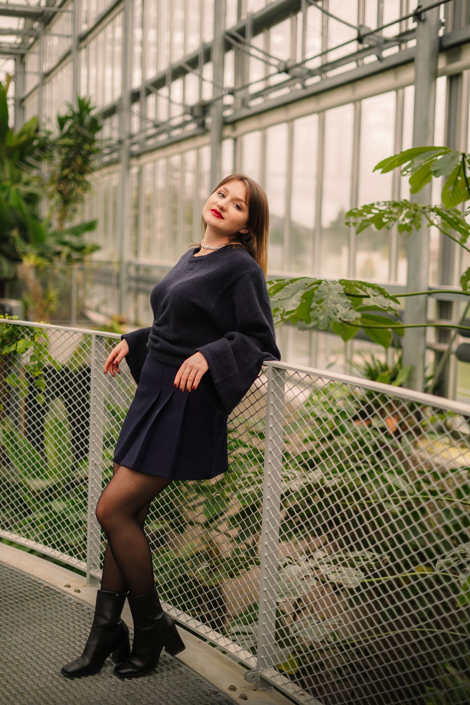 Elegant woman in navy blue dress posing in a greenhouse with tropical plants.