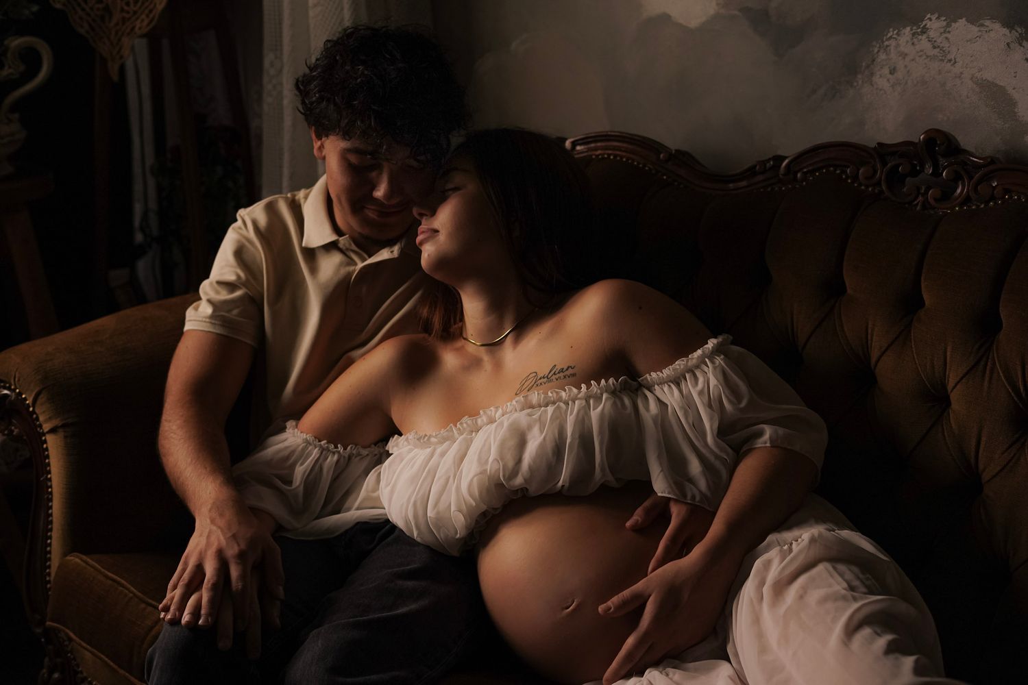 Intimate maternity photo of pregnant couple resting on vintage velvet sofa in moody studio lighting.