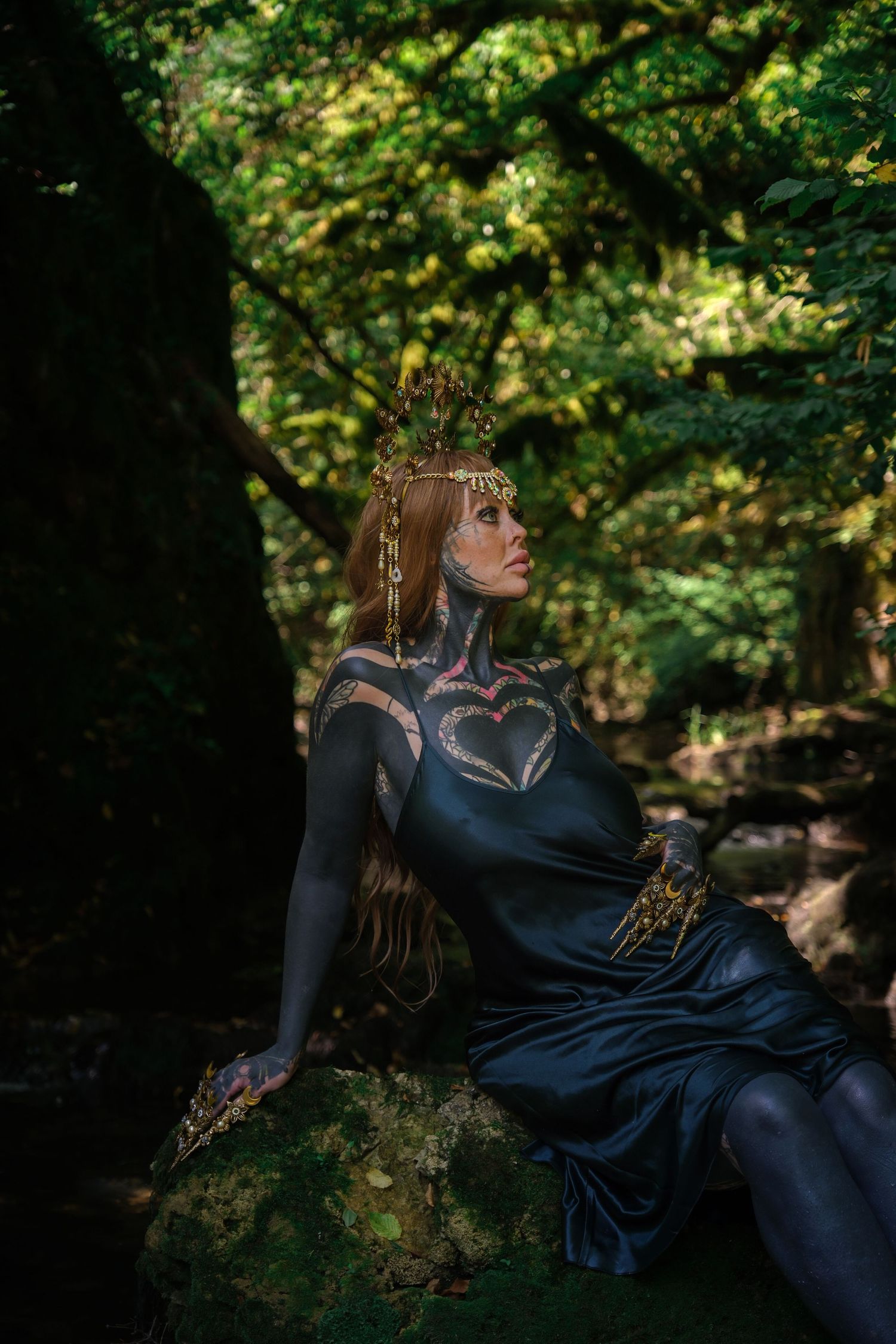 Woman in black bodysuit with body art sits on mossy rock in dark forest wearing ornate gold crown.