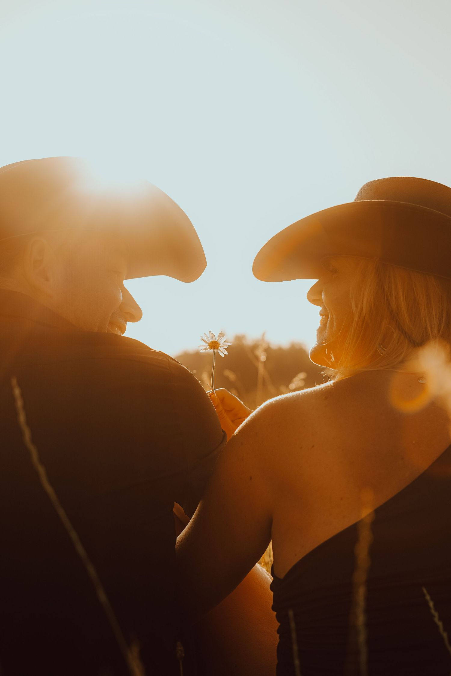A couple in cowboy hats face each other holding a daisy, bathed in warm golden sunset lens flare.