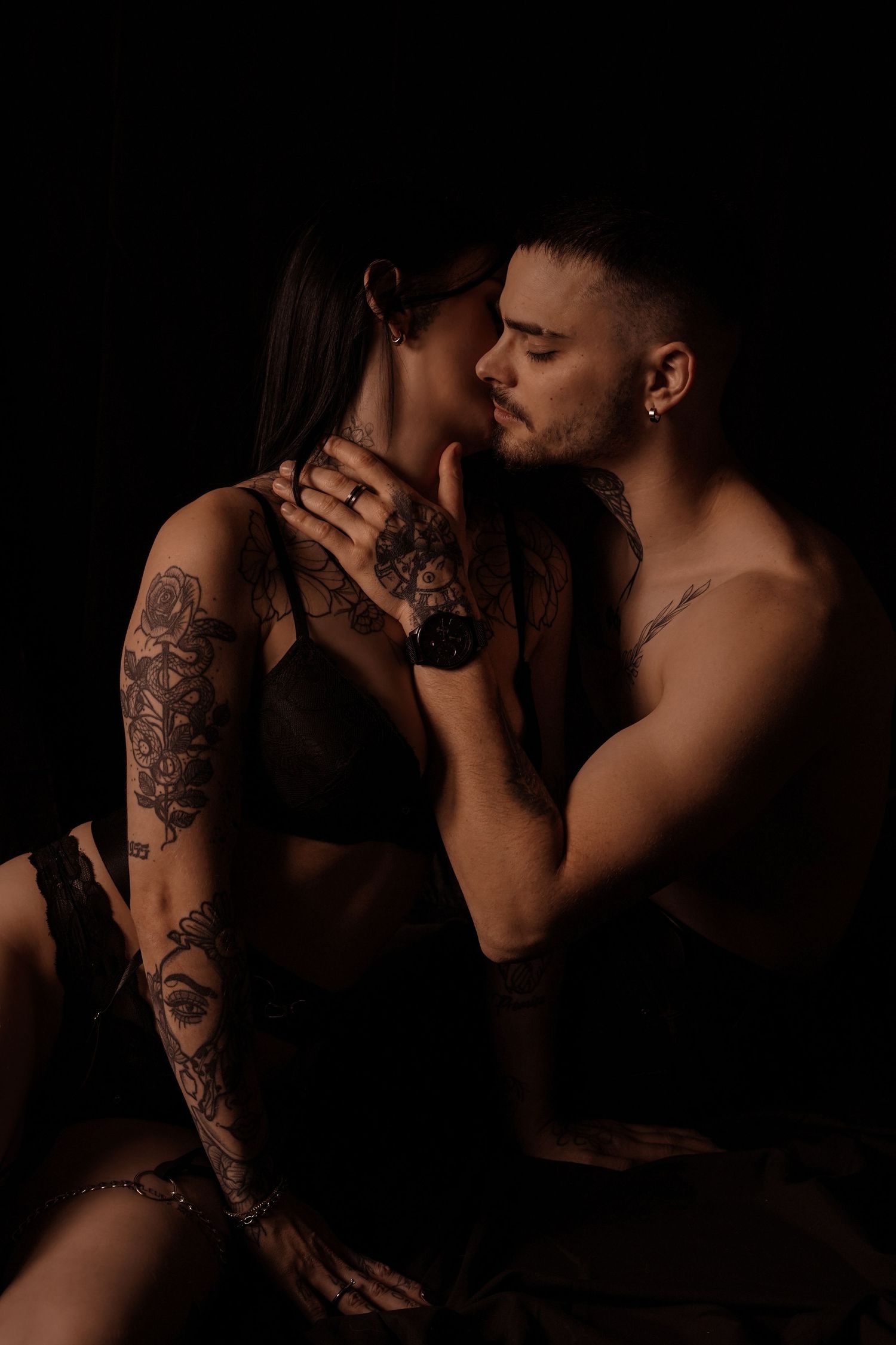 Tattooed couple in intimate embrace against dark background, man tenderly touching woman's face.