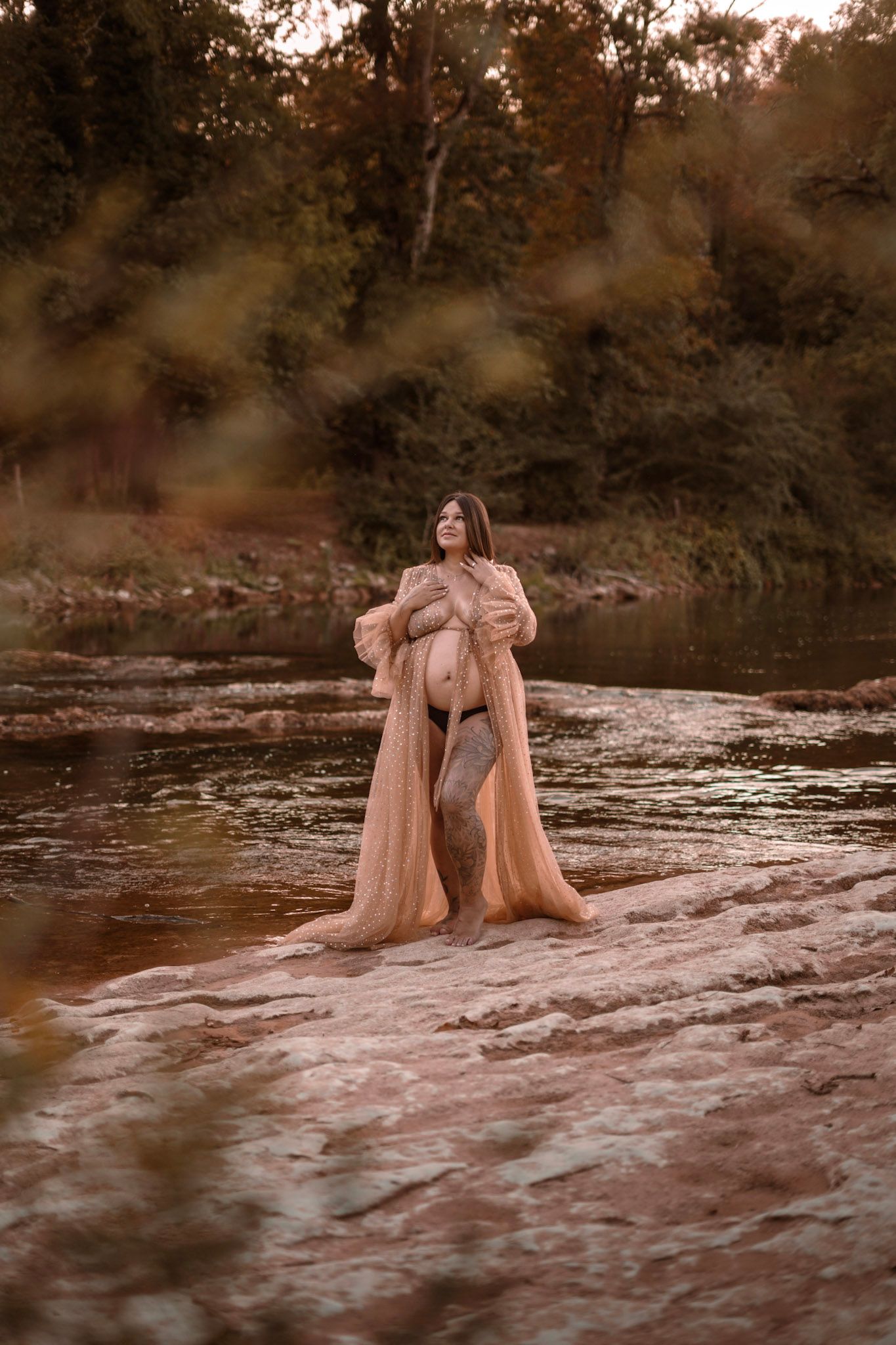 Pregnant woman in sheer peach robe standing on rocks by a river surrounded by autumn trees.