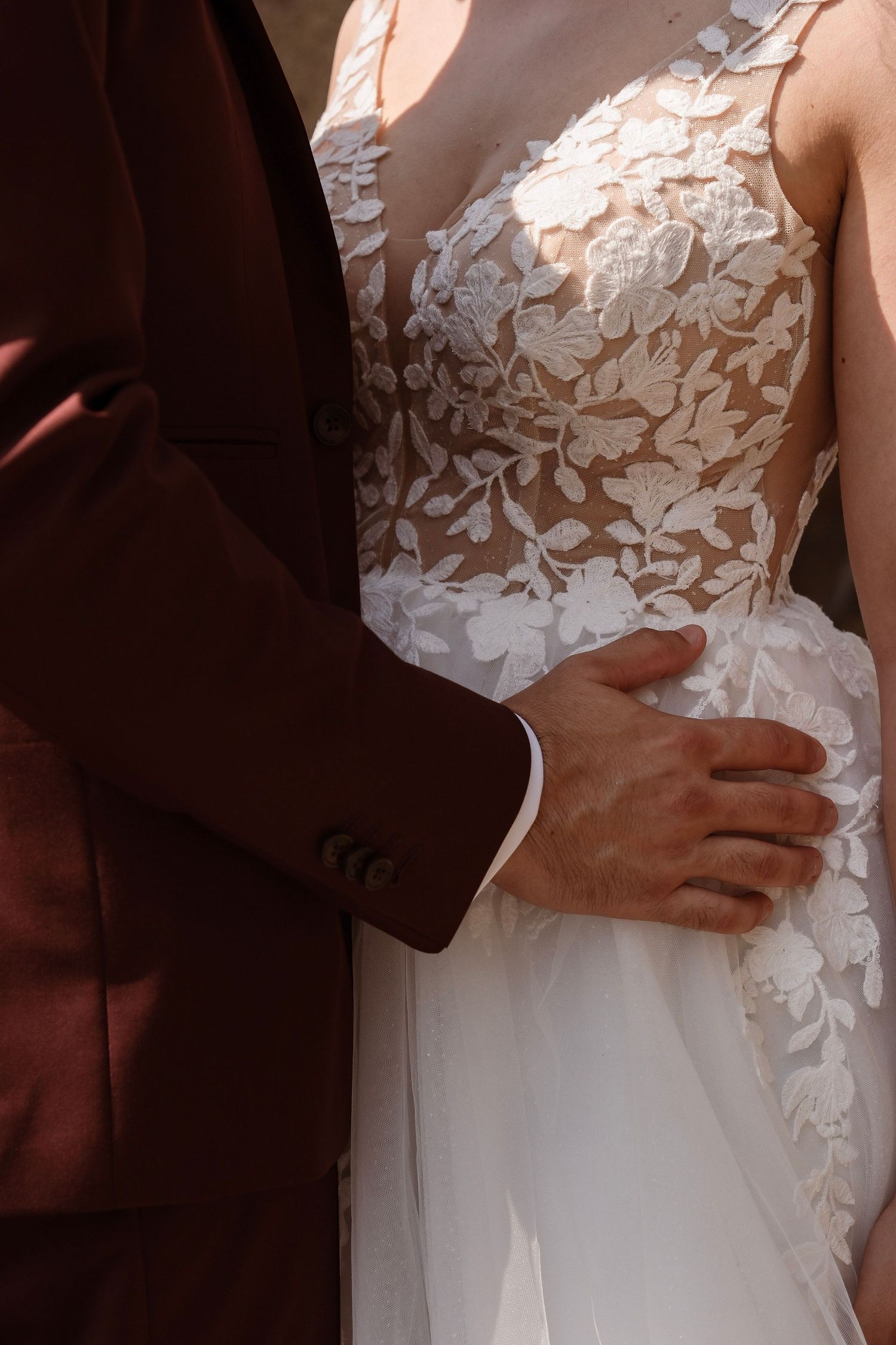 Close-up of groom in burgundy suit placing his hand on bride's waist in a detailed lace wedding gown.
