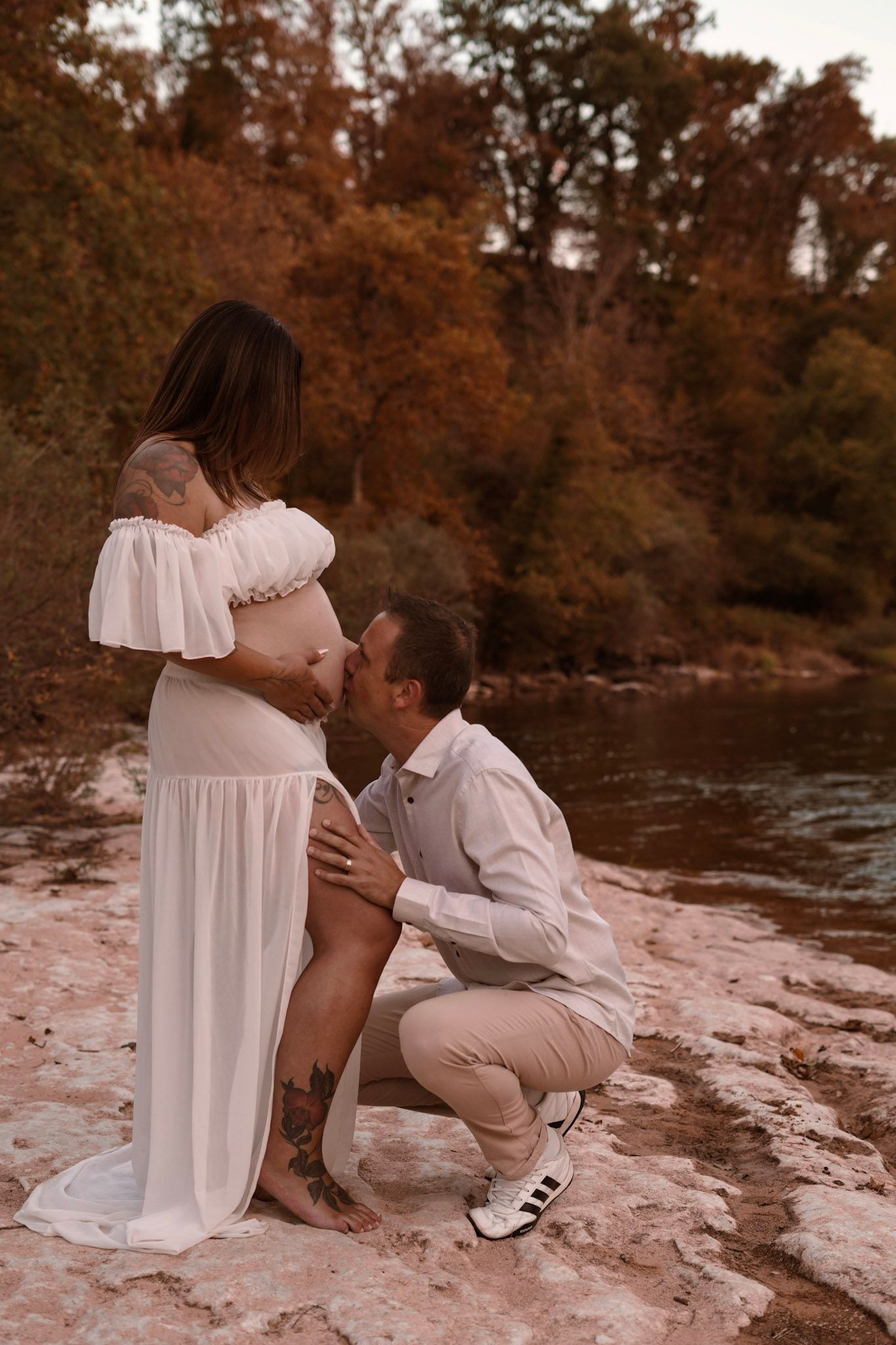 Man kneeling to kiss pregnant woman's belly outdoors near river with autumn foliage.