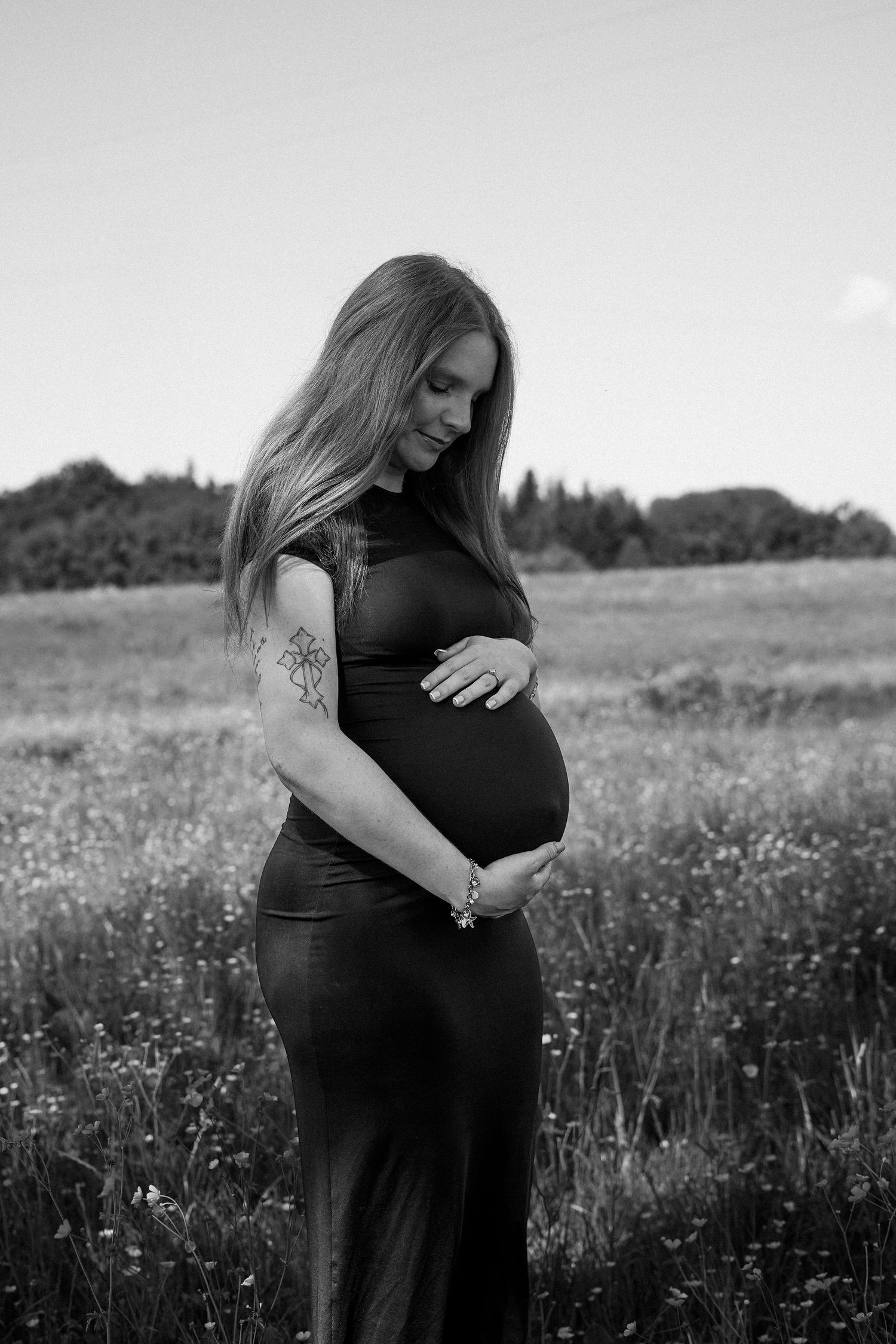 Black and white portrait of pregnant woman in dark dress cradling baby bump in field.