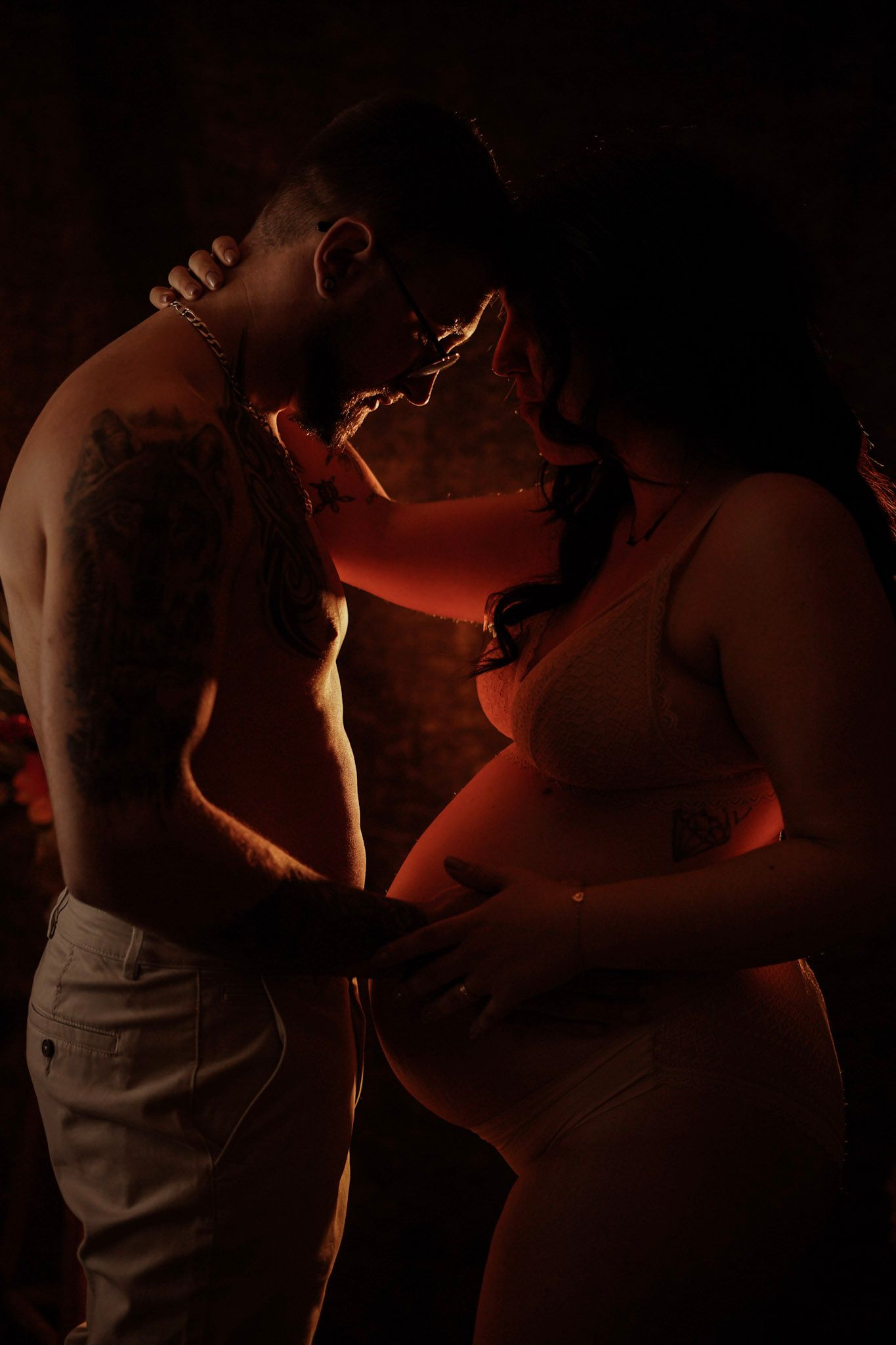 A shirtless tattooed man and pregnant woman share an intimate moment in warm red-toned studio lighting.