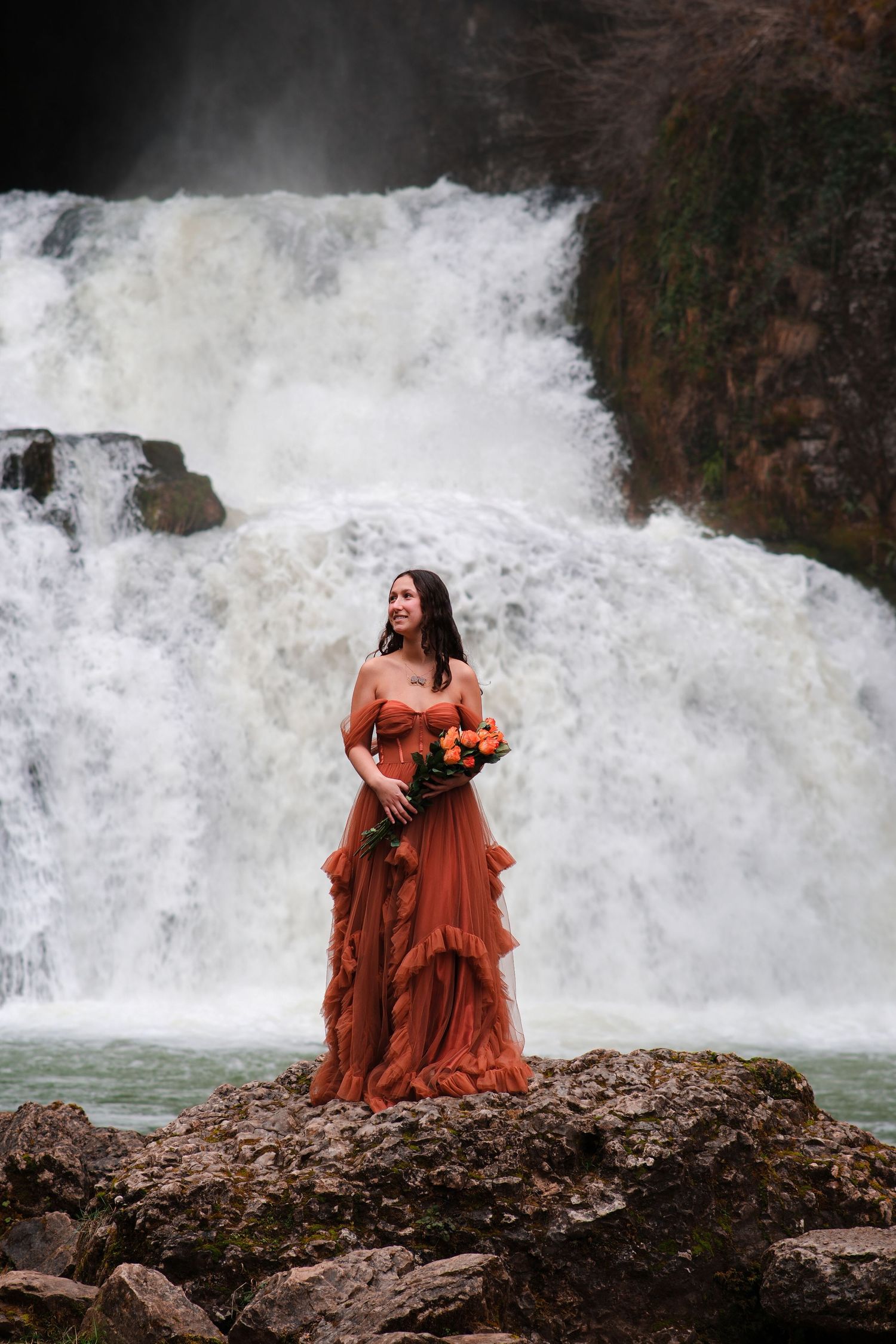 Woman in rust orange ruffled gown holds bouquet standing on rocks before dramatic rushing waterfall.