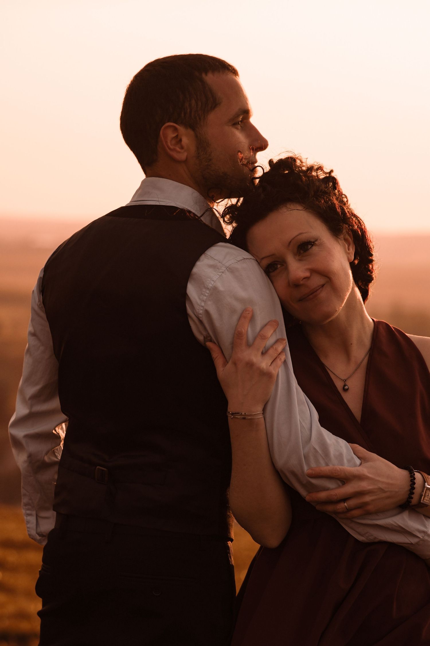 A couple embraces at sunset, woman resting her head on mans chest in a warm golden field.