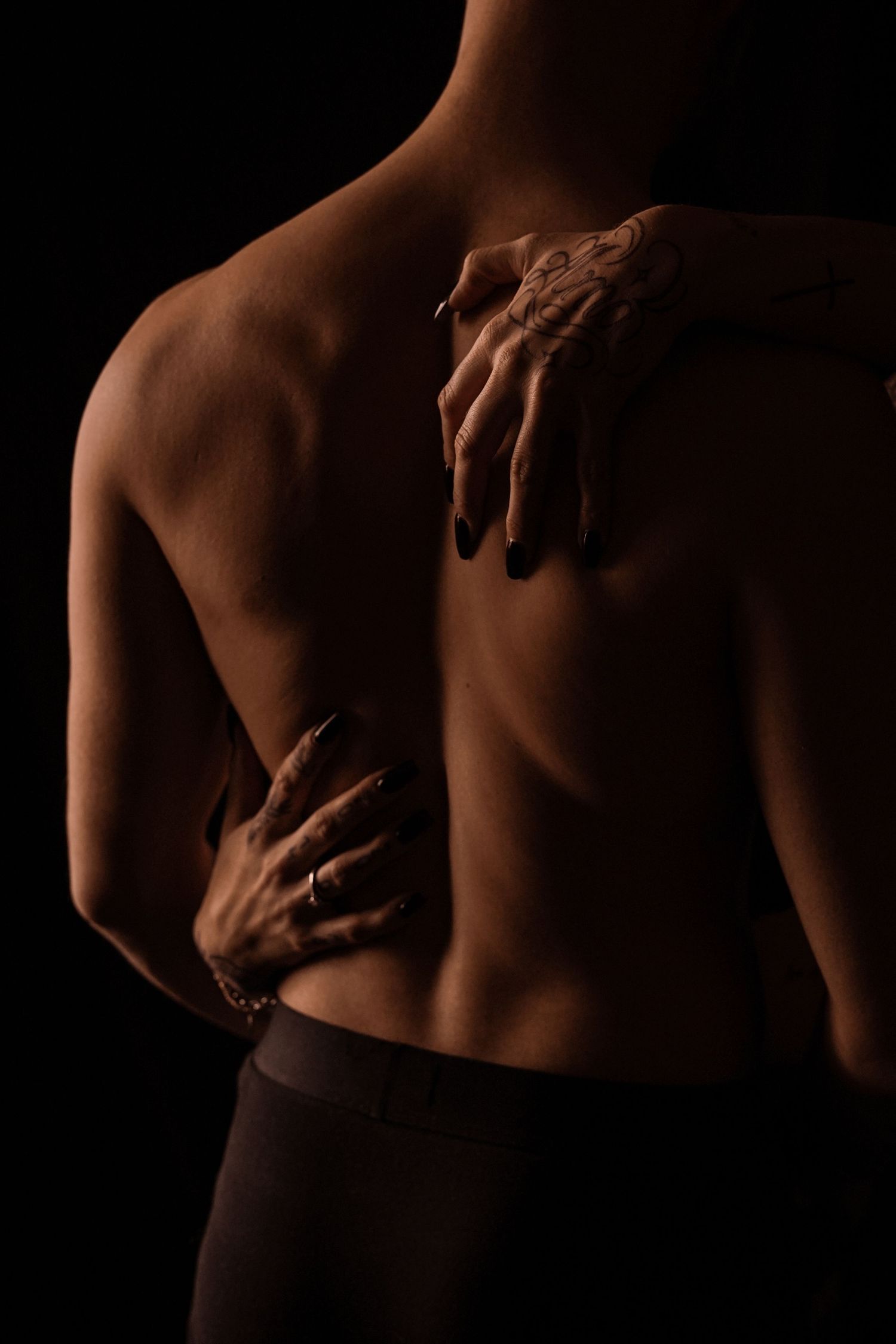 Woman's hands gently placed on shirtless man's muscular back in dark moody photography.