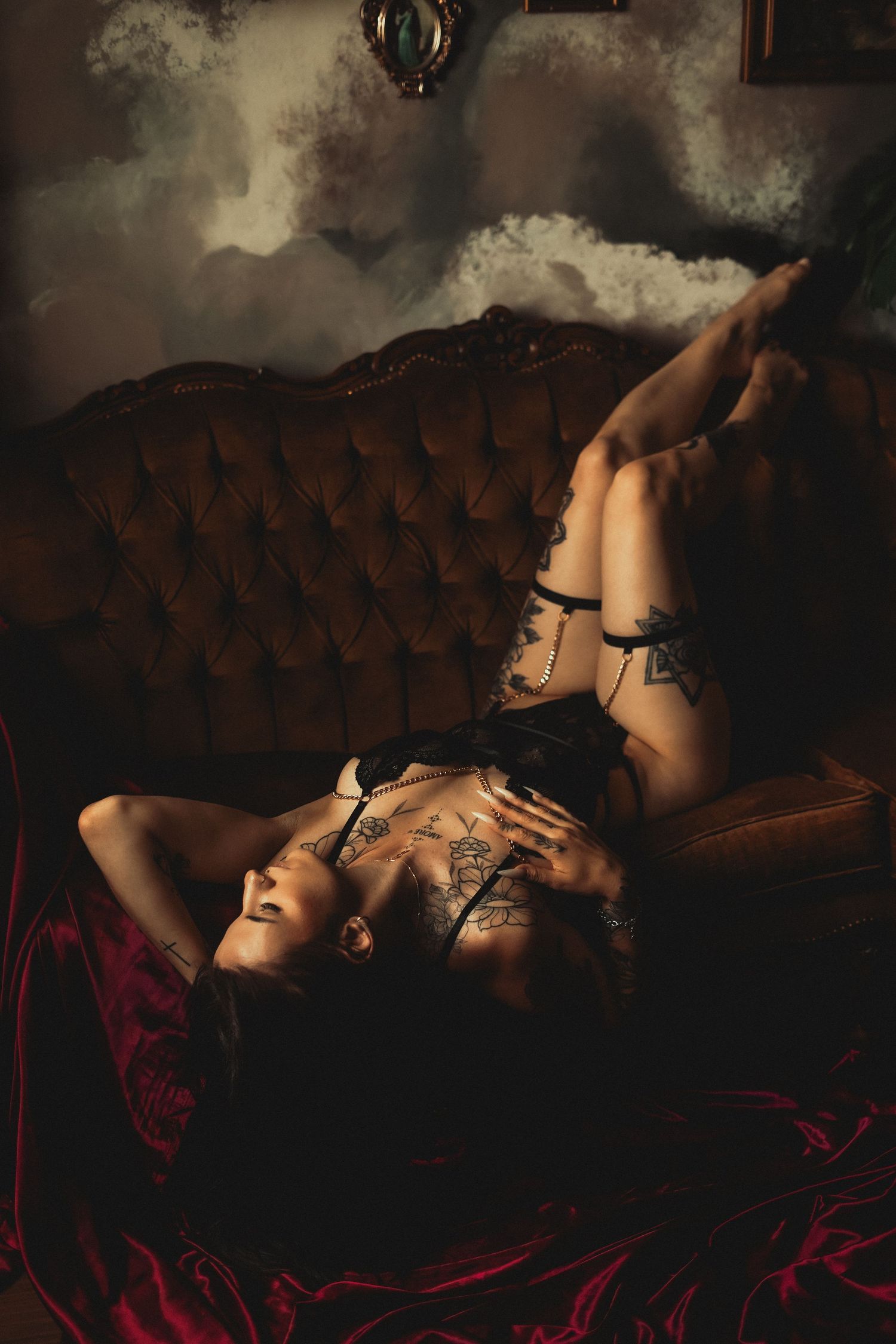 Tattooed woman in black lingerie lying on vintage velvet sofa with dramatic moody lighting.