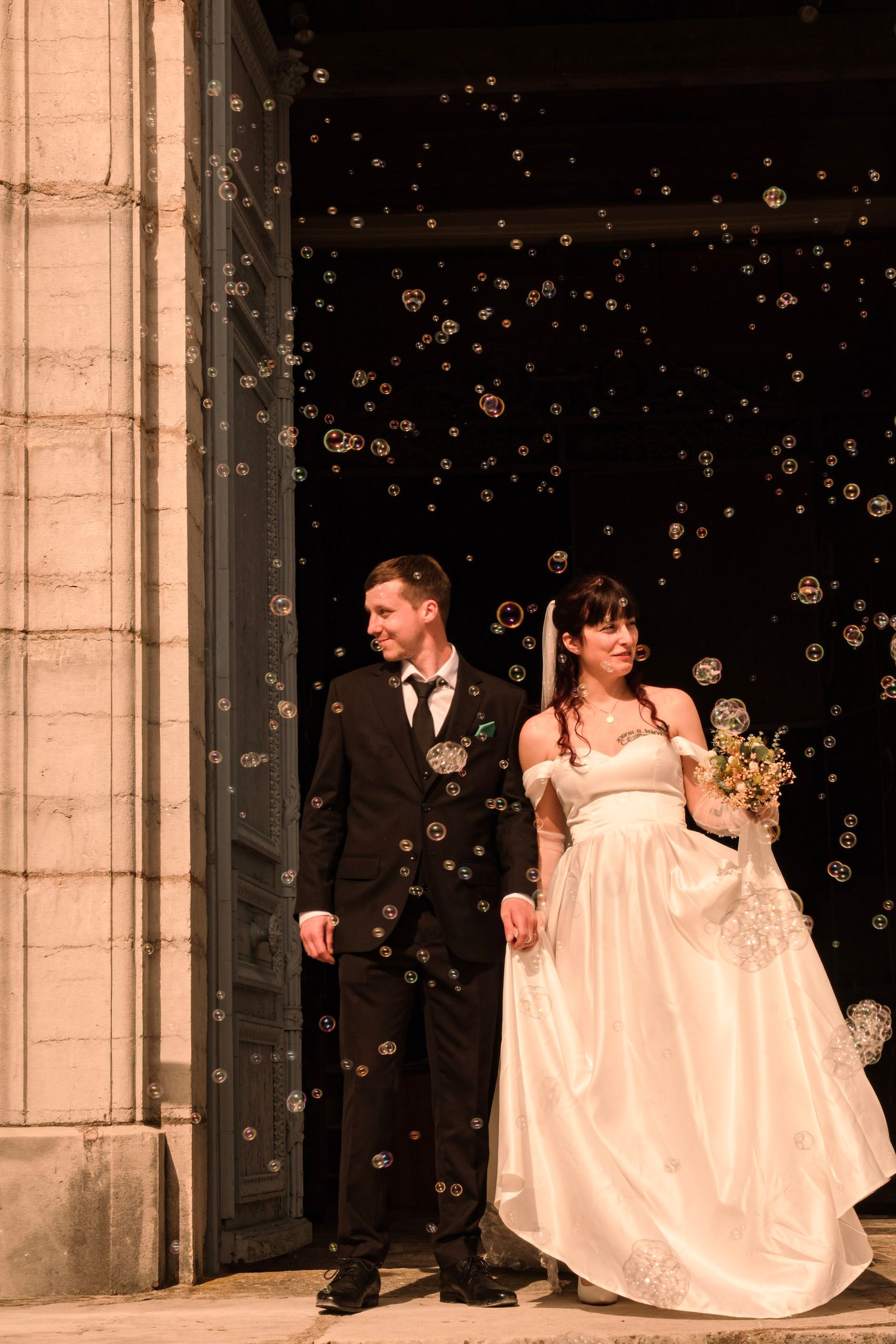 Newlyweds exit a grand church doorway surrounded by a shower of floating bubbles at night.