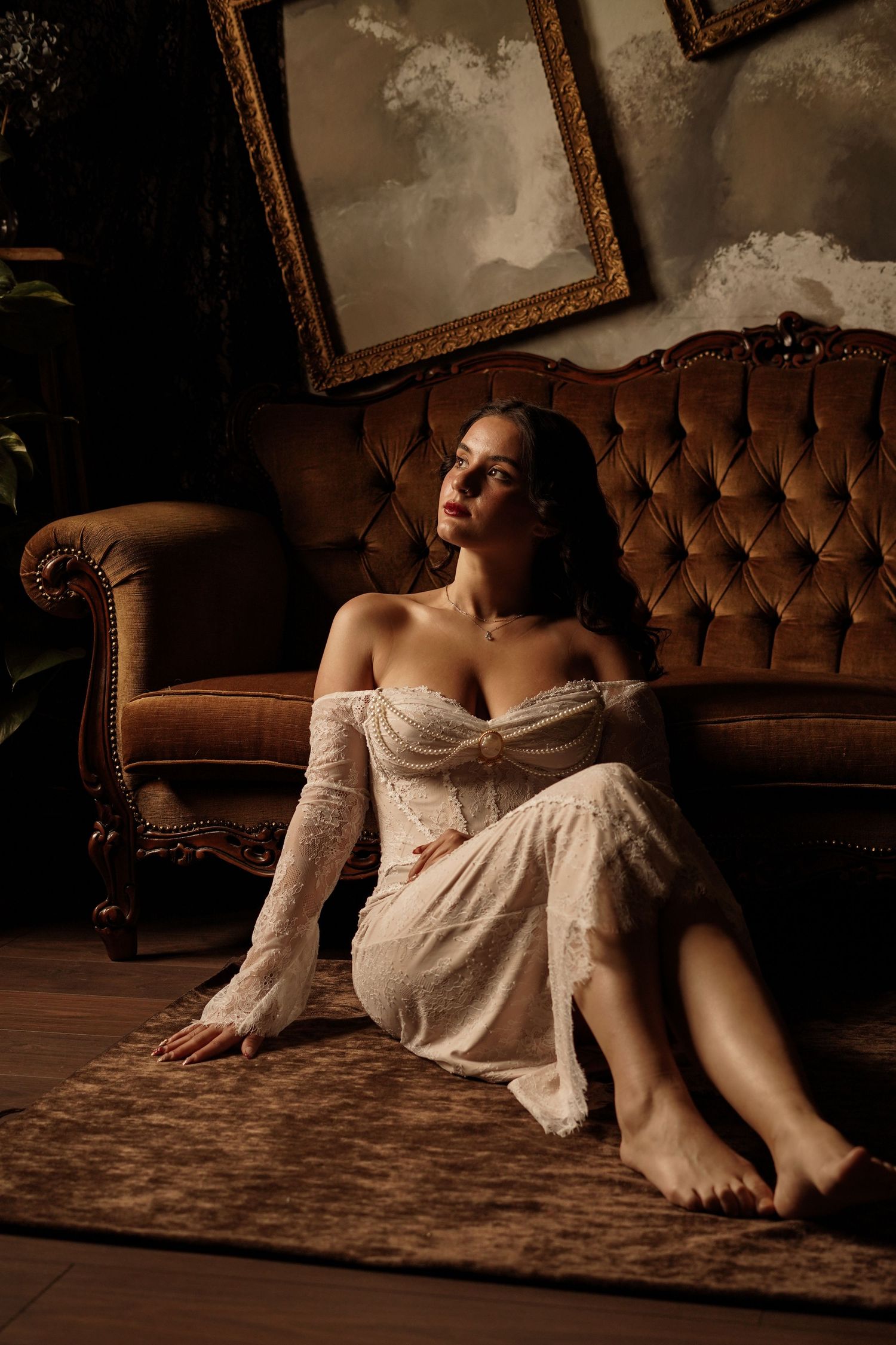 Brunette woman in white off-shoulder lace dress sitting on floor beside vintage tufted sofa.