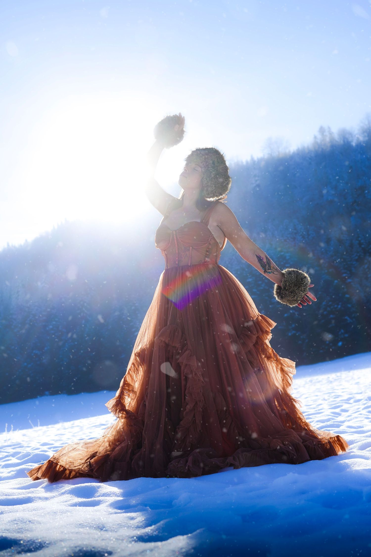 A woman in a flowing rust gown stands in snowy mountains with arms raised in bright winter sunlight.