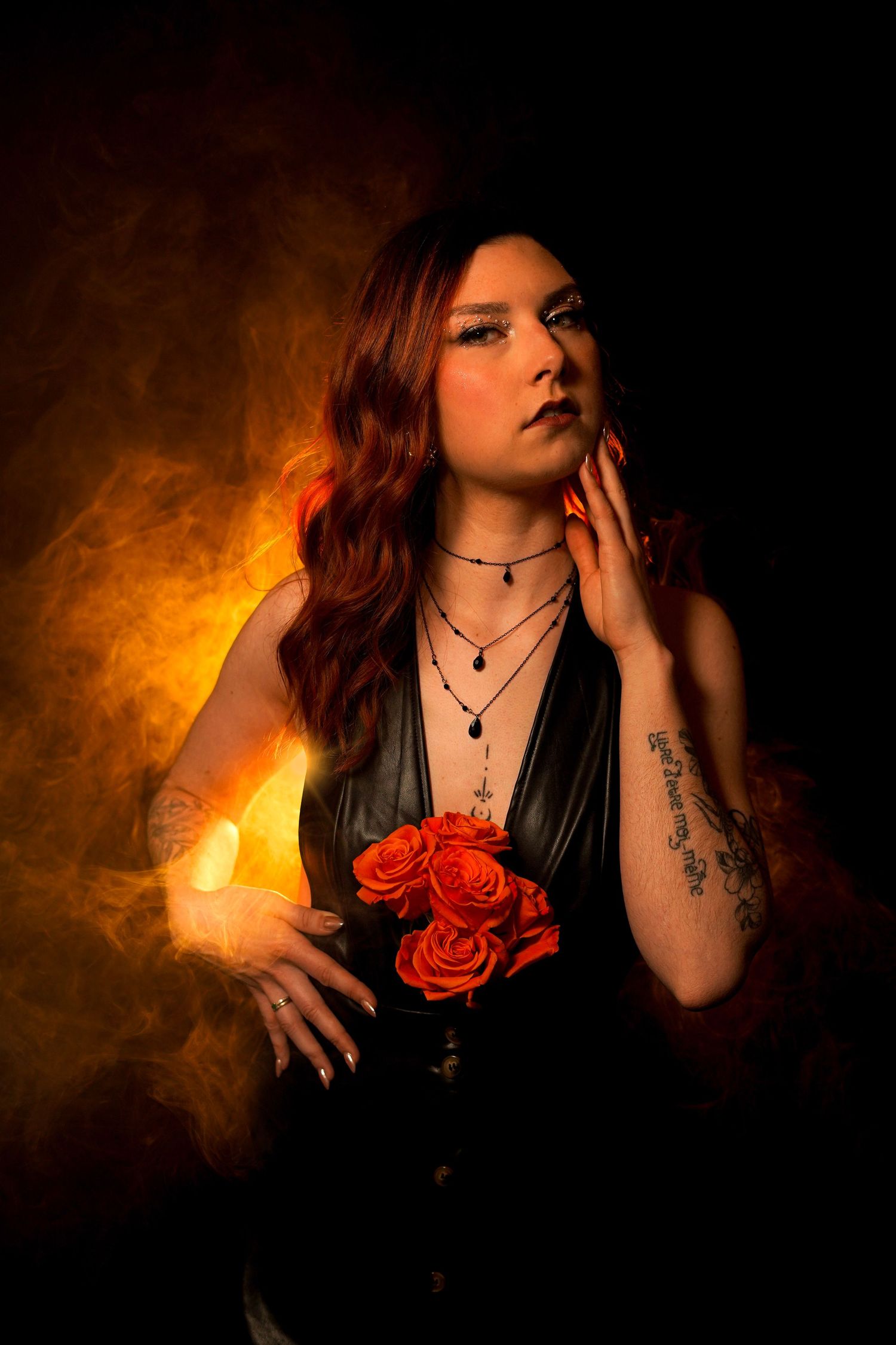 Dramatic portrait of redhead woman in black dress holding red roses surrounded by orange smoke.