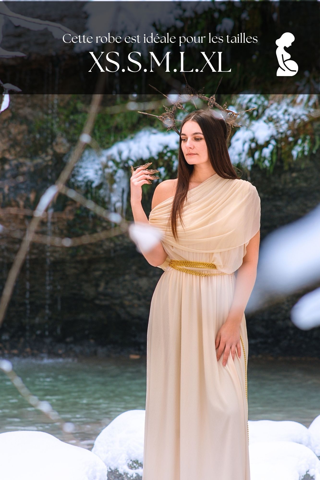 Woman in elegant cream Greek-style gown with gold belt, wearing floral crown, posing in snowy winter forest setting.