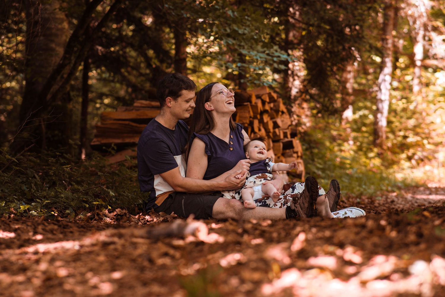 Family sitting on autumn forest floor with baby, surrounded by fallen leaves and warm golden light.