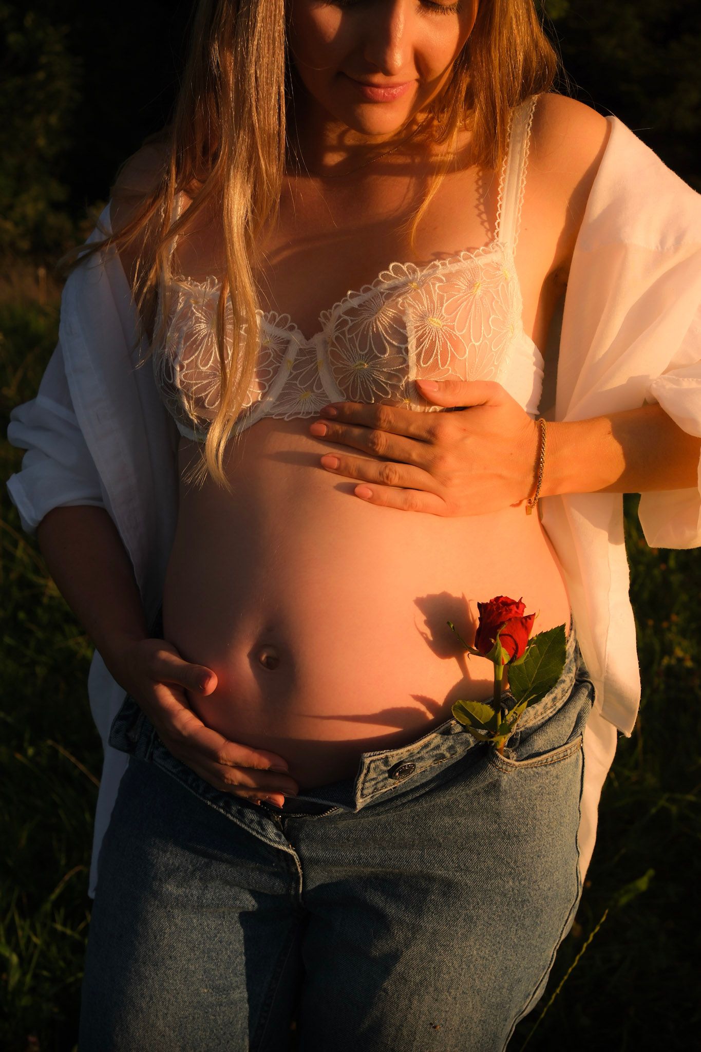 Pregnant woman in white lace bra and jeans holding a red rose against her baby bump in golden sunset light.