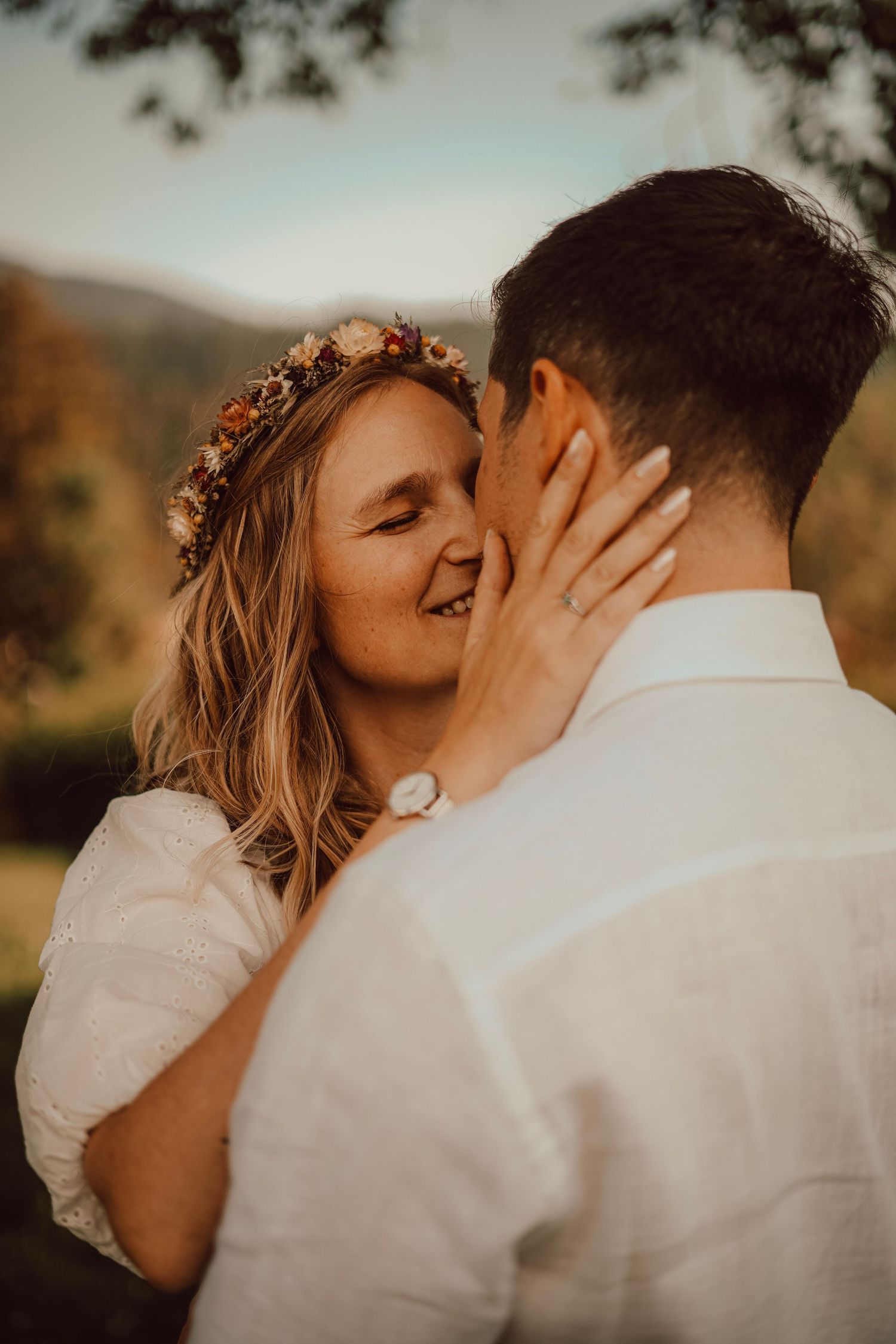 Smiling bride wearing a floral crown tenderly holds grooms face during outdoor wedding portrait.