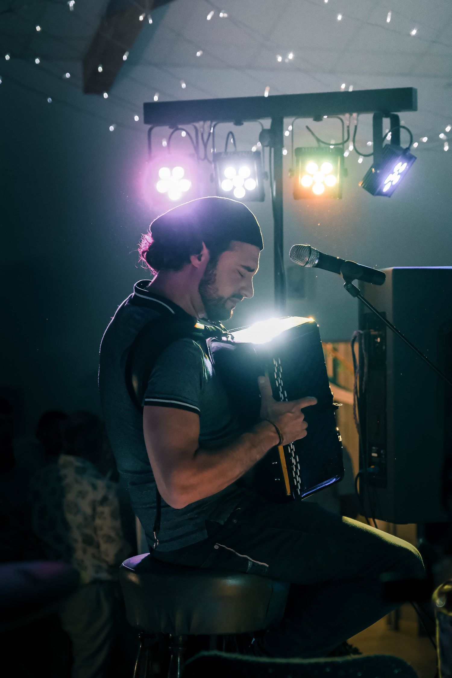 Male accordion player performing on stage under colorful stage lights during indoor live music event.