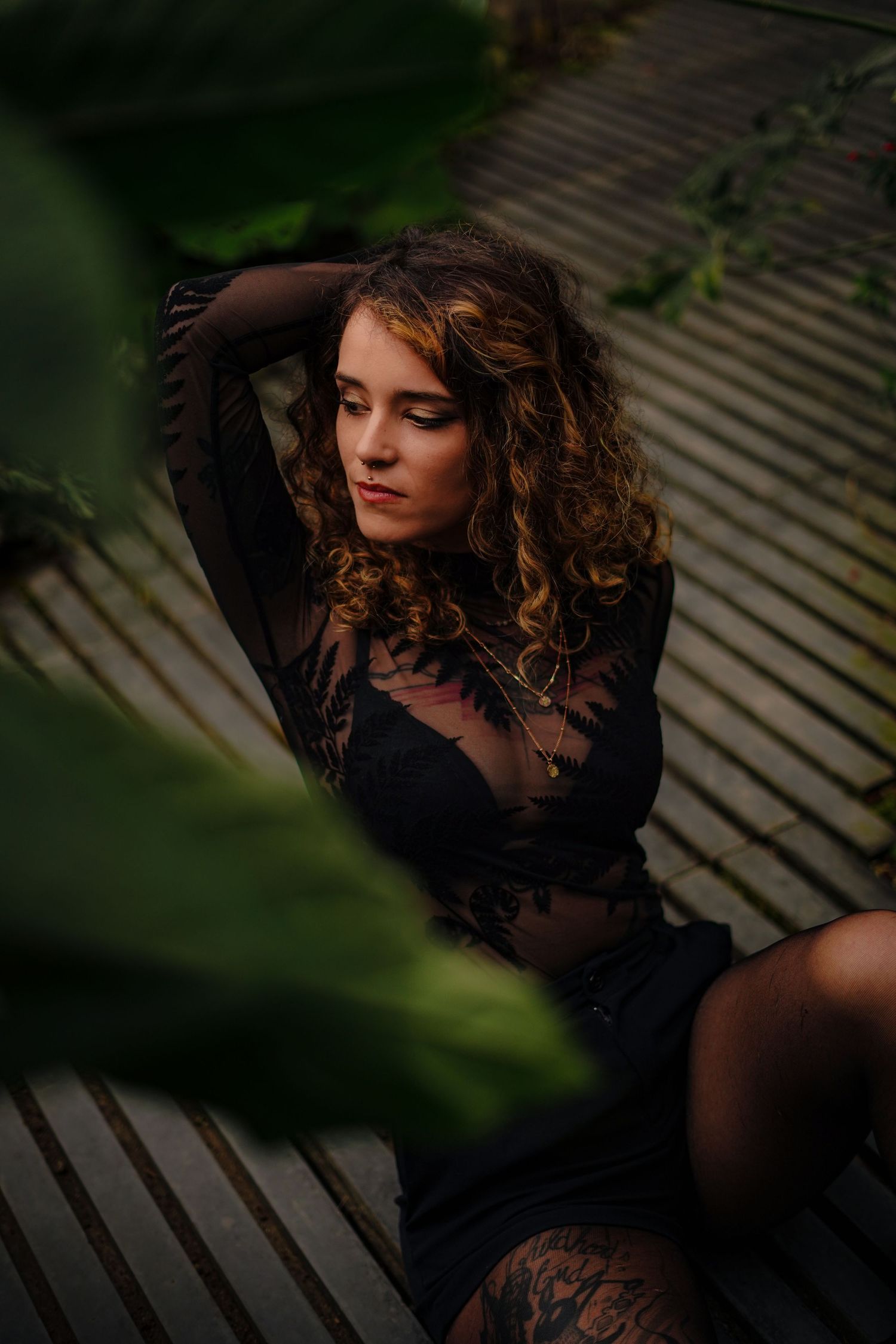 Curly-haired woman in black lace outfit sitting on wooden deck surrounded by foliage.
